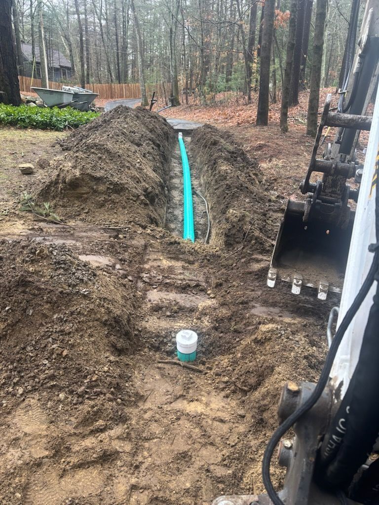 Trench with blue pipe installed, a white coupling, and a bobcat machine, in a wooded outdoor setting.