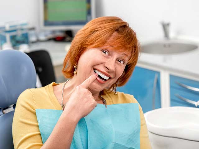 A woman is sitting in a dental chair and pointing at her teeth.
