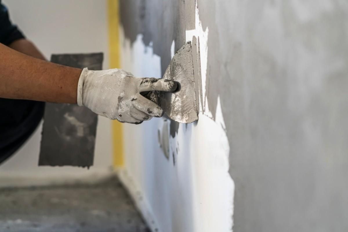 Gloved hand spreading white plaster on a gray wall with a trowel