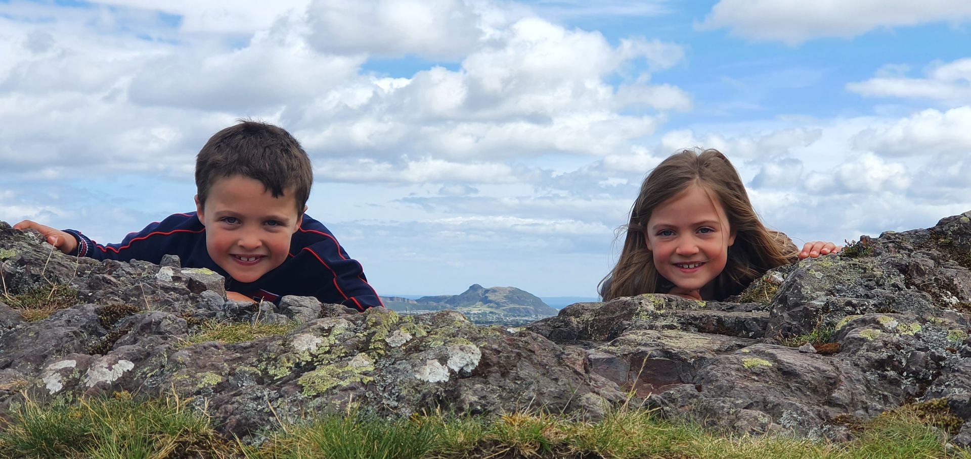 View of Arthurs Seat in Edinburgh, as seen from Torduff Hill, Pentlands Regional Park. This is in the background and in between two children looking over a rocky outcrop. Sky is blue.