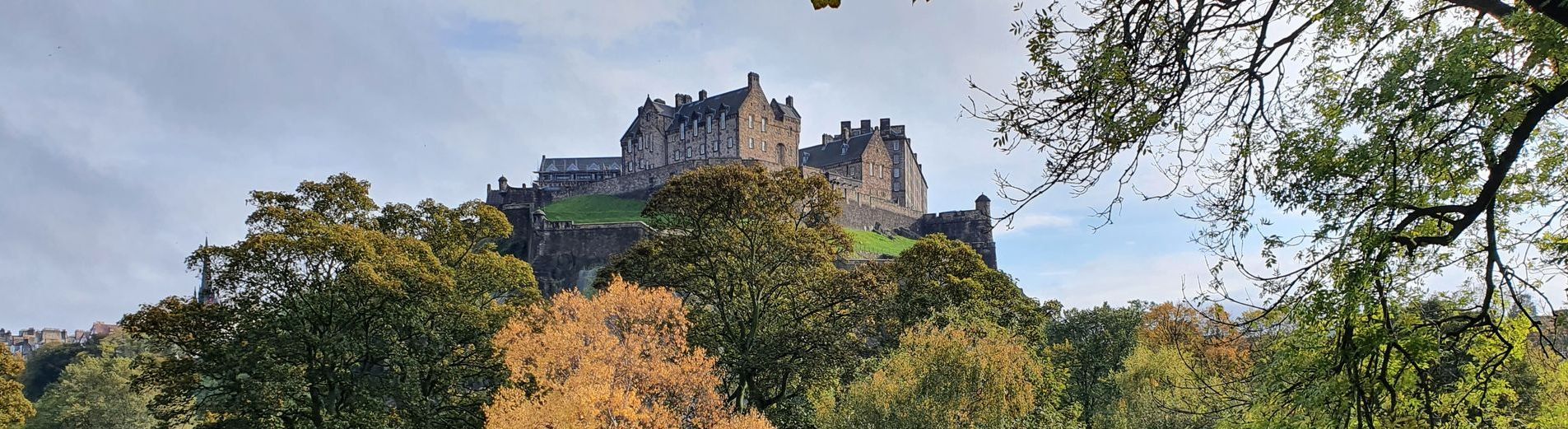 Image of Edinburgh Castle on a sunny day with trees from Princes Street Gardens (West) in the foreground. Image belongs to Tartan Compass Tours.