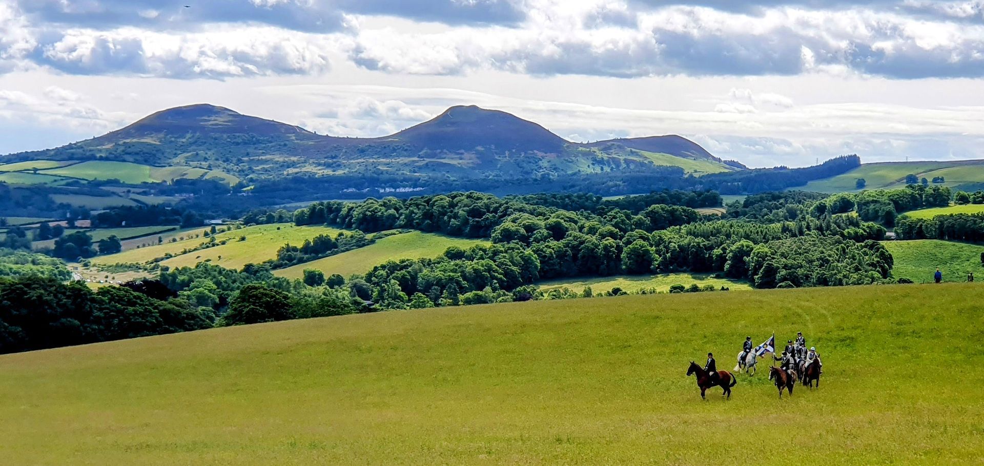 Image of horse riders carrying a flag standard whilst doing their ride out on gala day. This is during the Braw Lads Gathering, Galashiels. They are in a meadow with expansive views behind them of farmland, woodland and the three eildon hills, near Melrose.