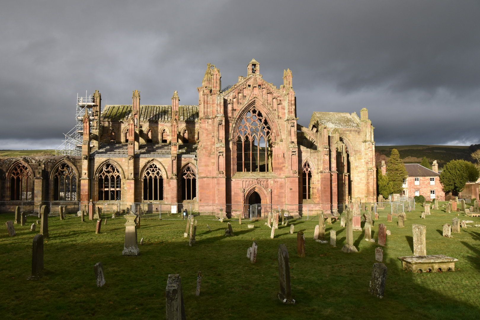 Image of the ancient ruin of Melrose Abbey in the Scottish Borders. Photo taken from the South side. An old graveyard, with damaged headstones, is in the foreground.