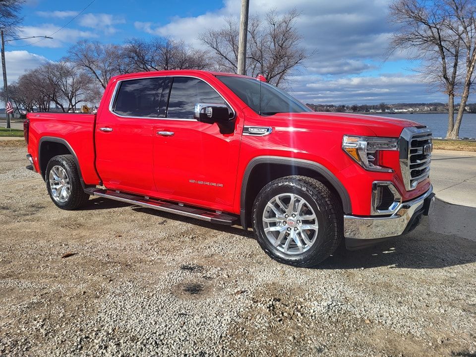 A red pickup truck is parked in a gravel lot next to a body of water.