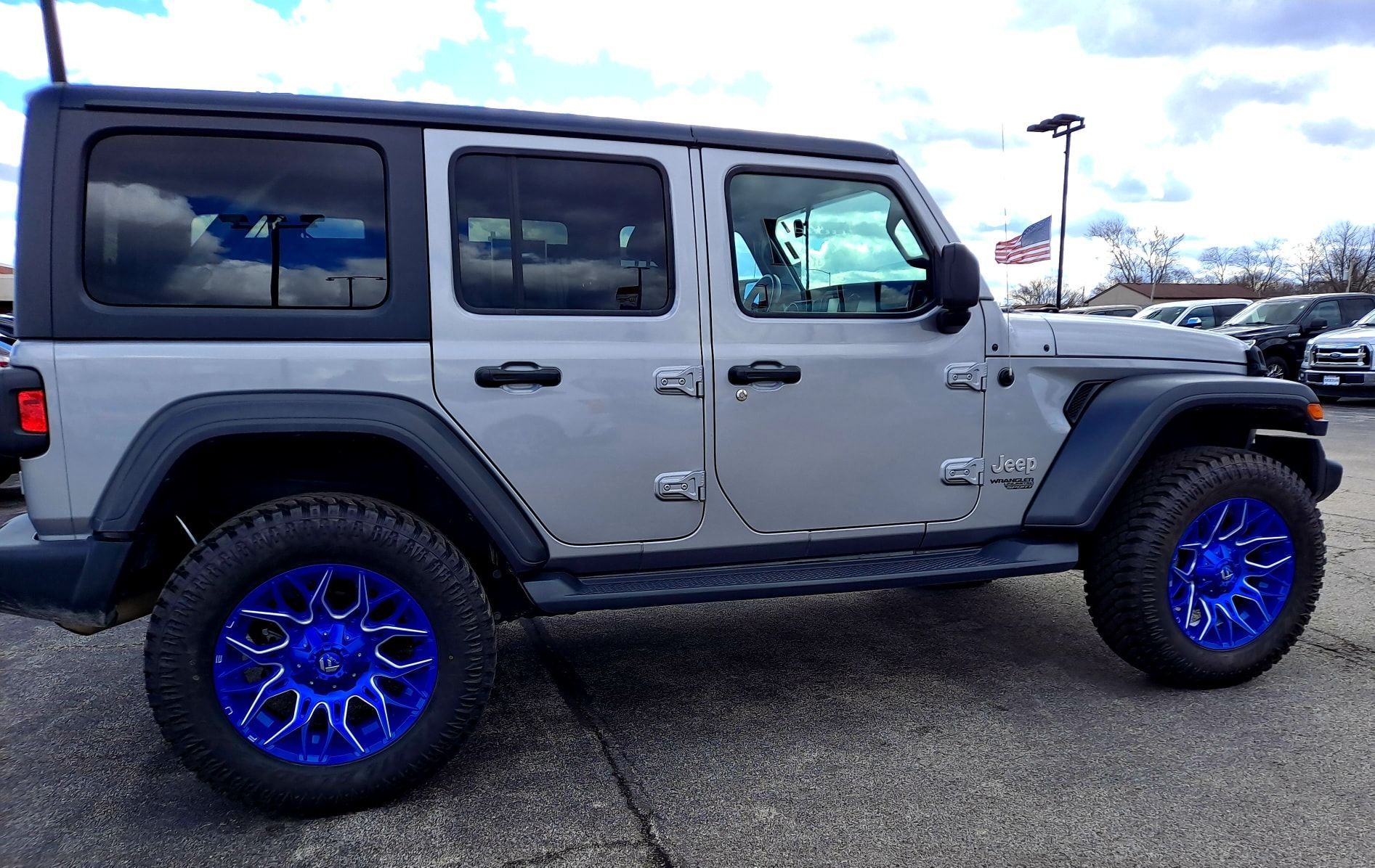 A silver jeep with blue wheels is parked in a parking lot.