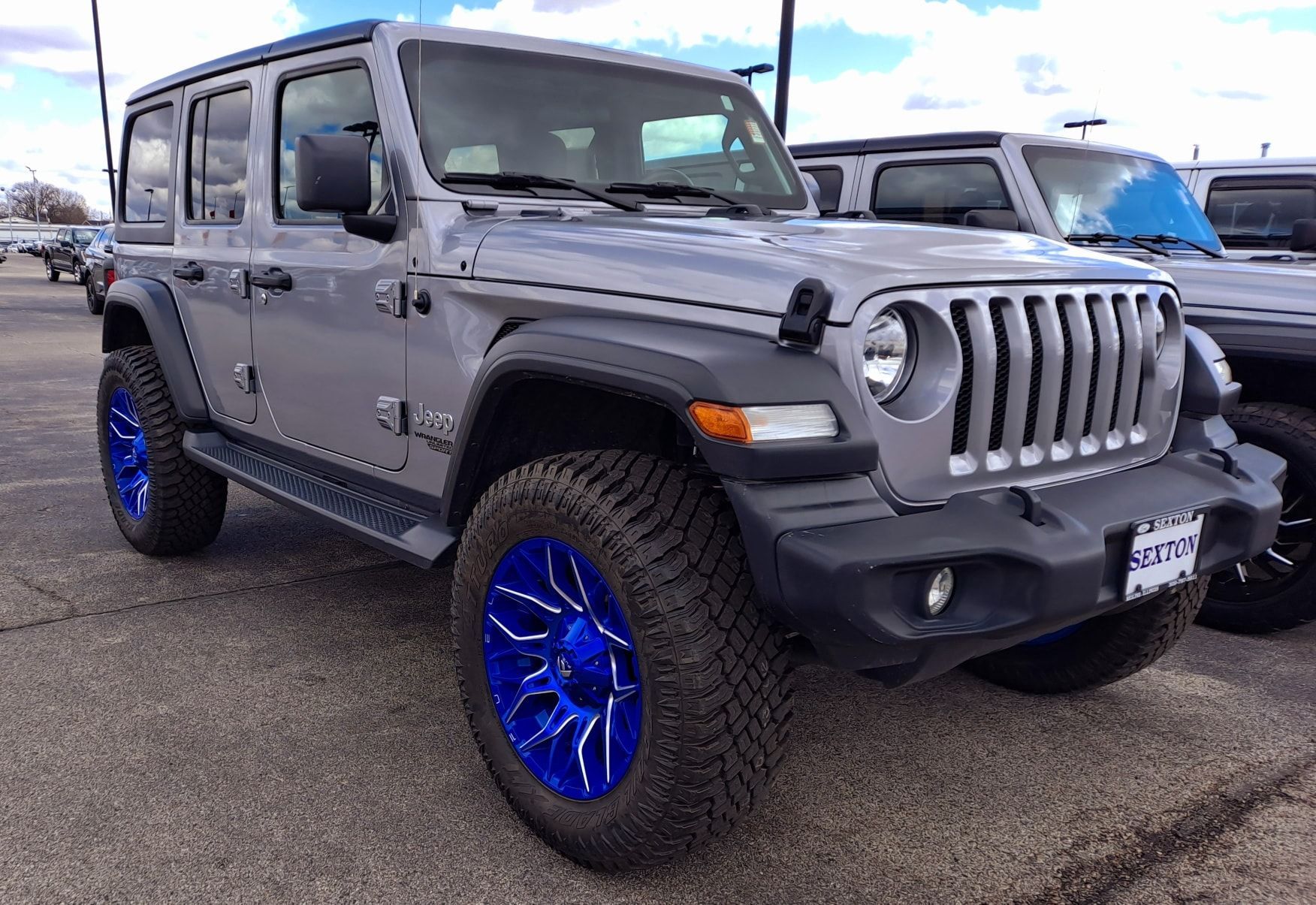 A gray jeep wrangler with blue wheels is parked in a parking lot.