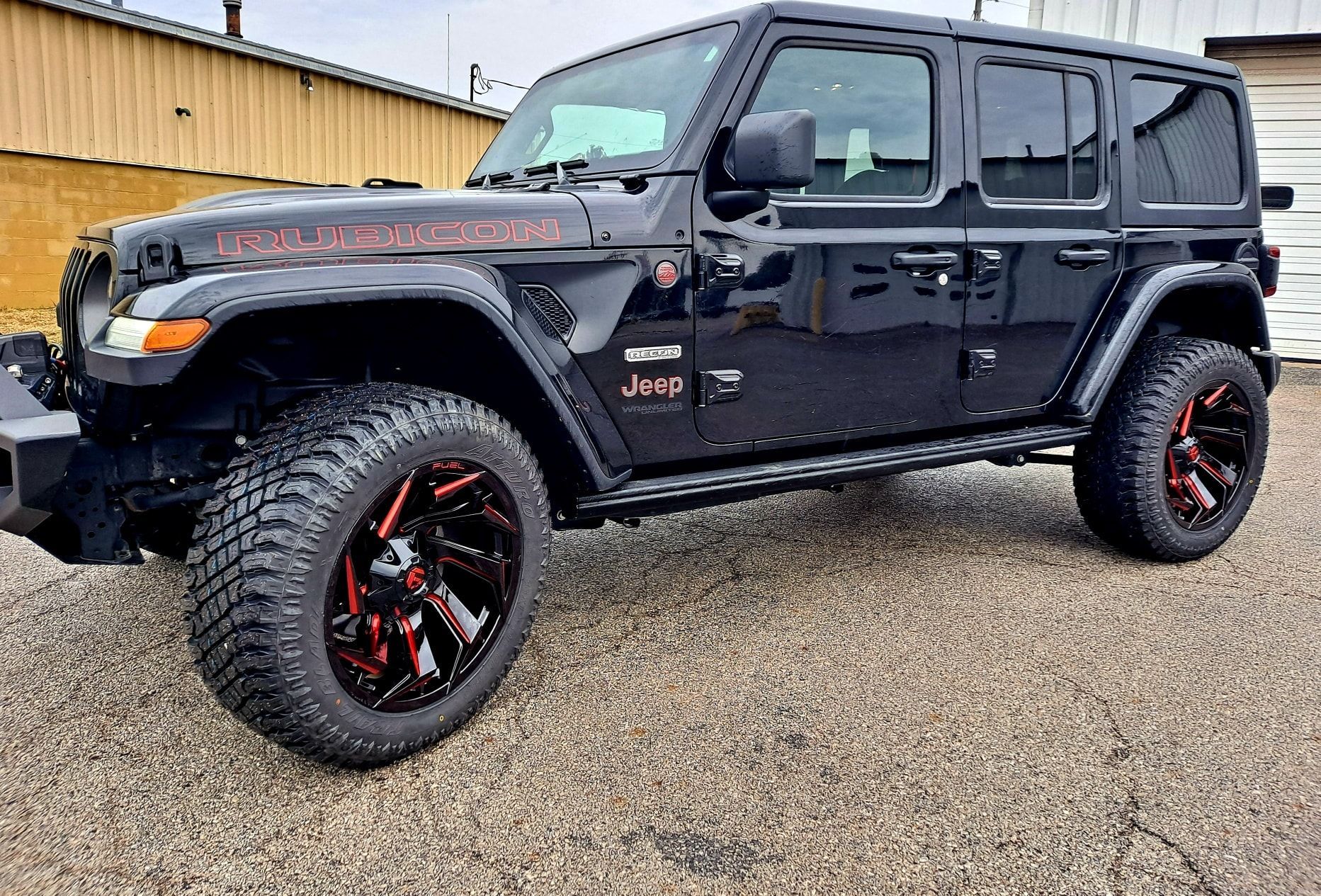 A black jeep wrangler with red wheels is parked in a gravel lot.