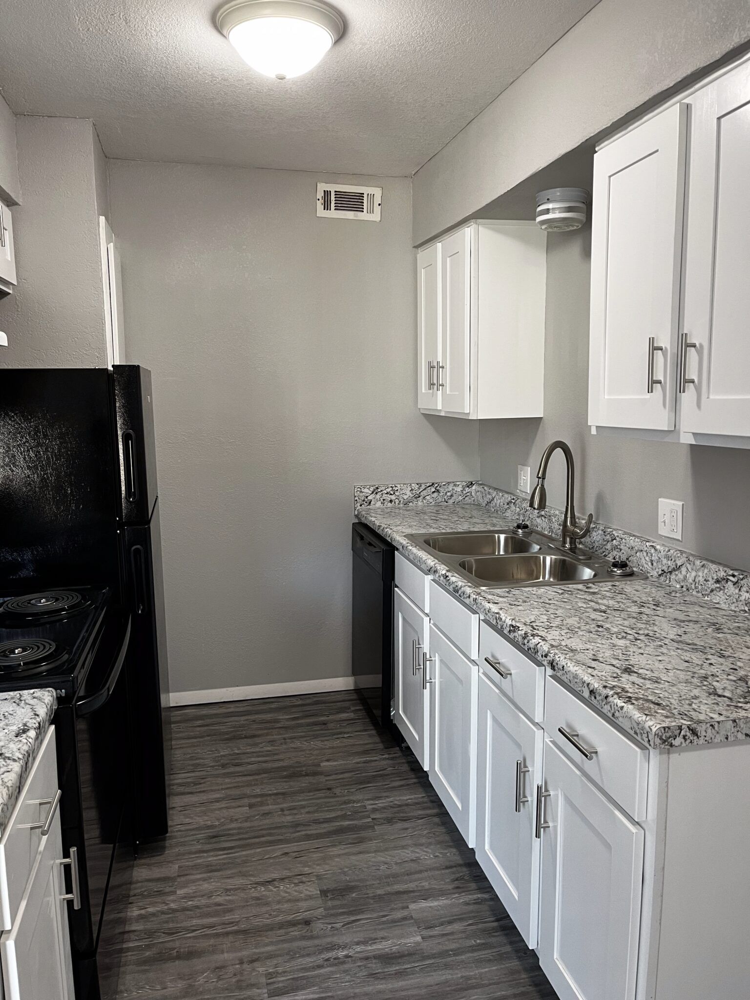 A narrow kitchen with white cabinets, gray walls, black appliances, and speckled countertops.