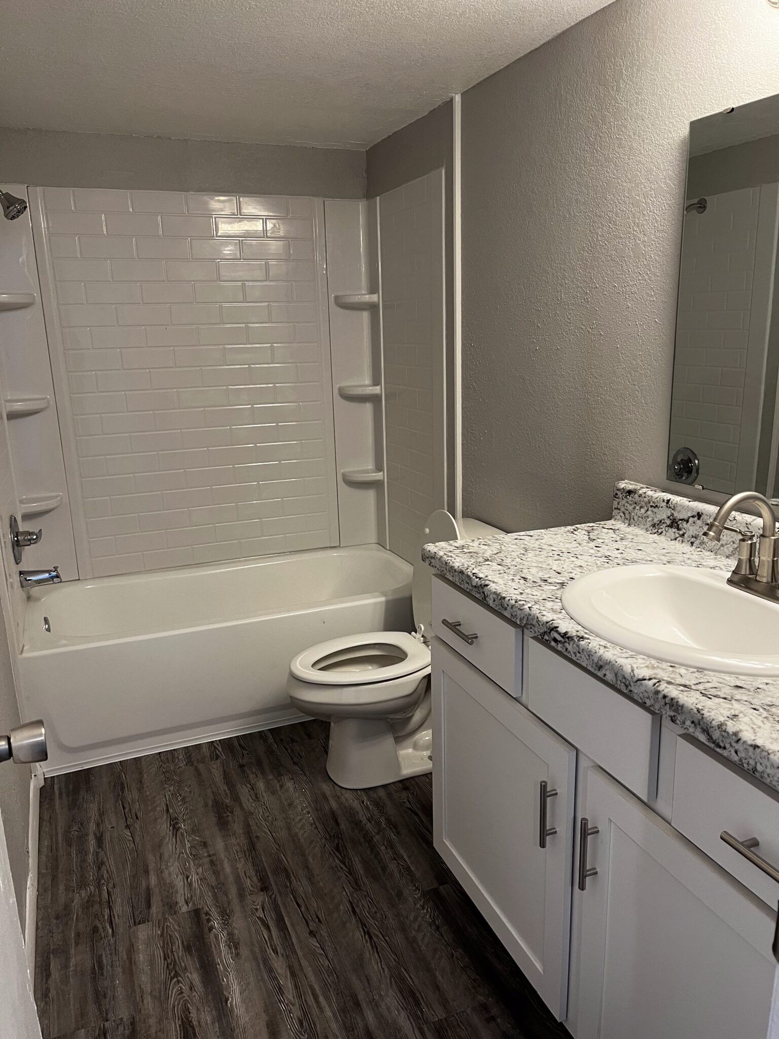 Bathroom with white bathtub, toilet, and vanity with speckled countertop; wood-look flooring.