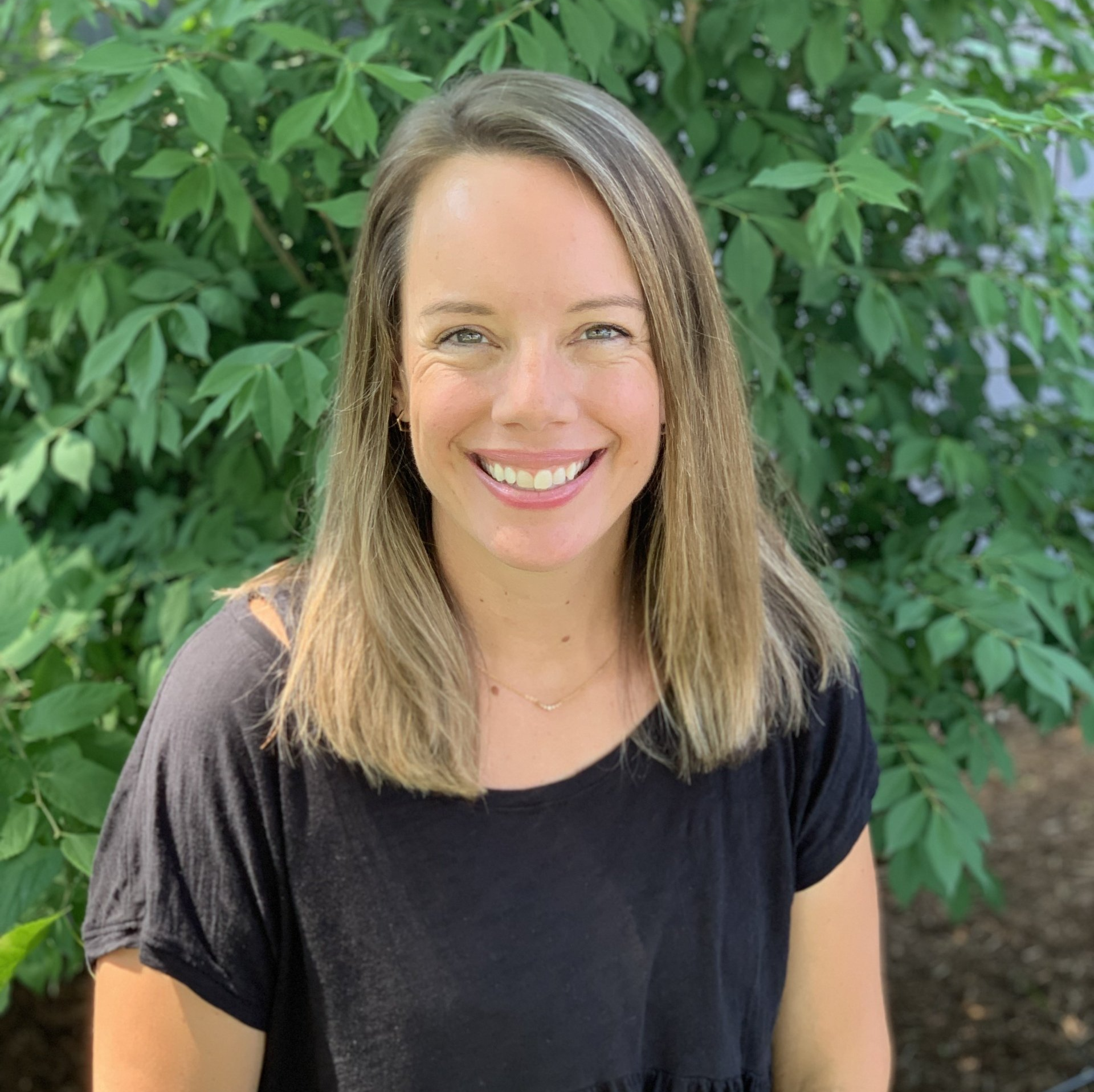 A woman in a black shirt is smiling in front of a tree.