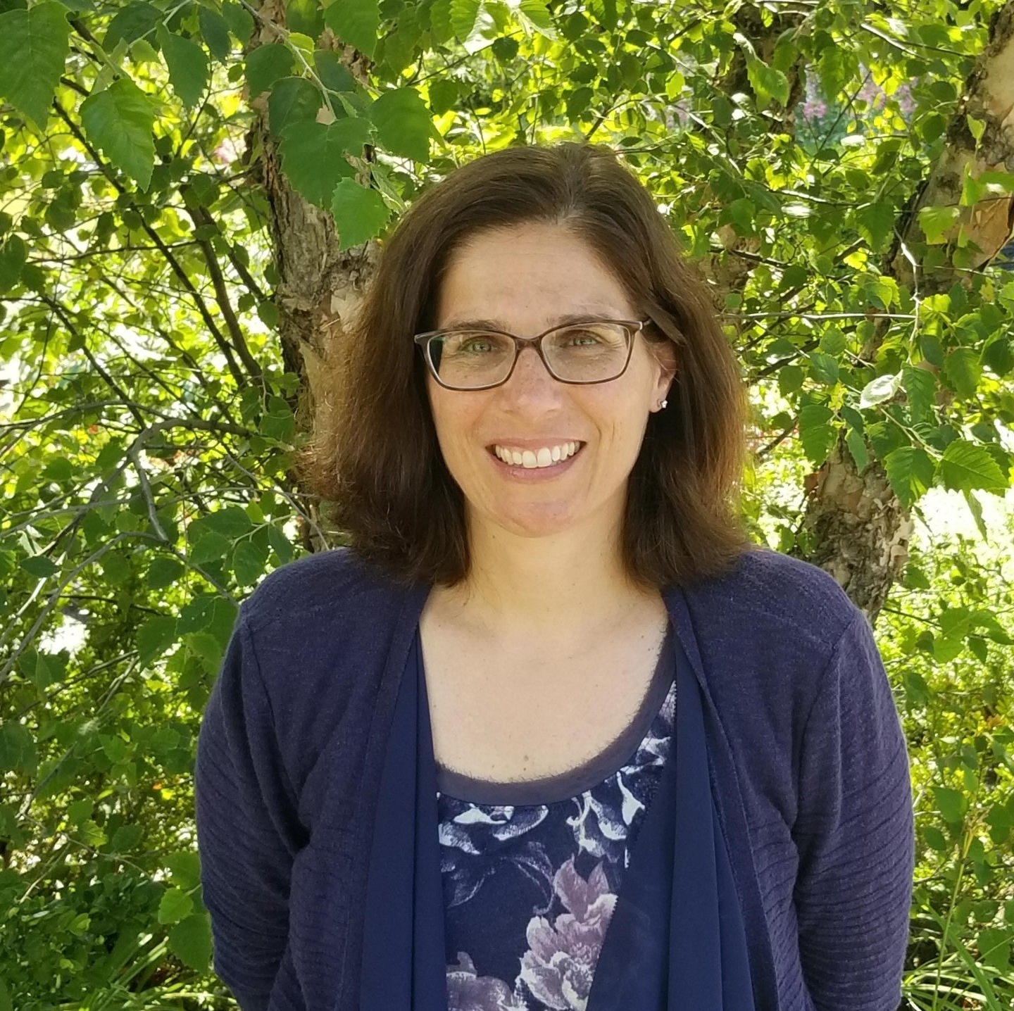 A woman wearing glasses and a blue sweater is smiling in front of a tree.