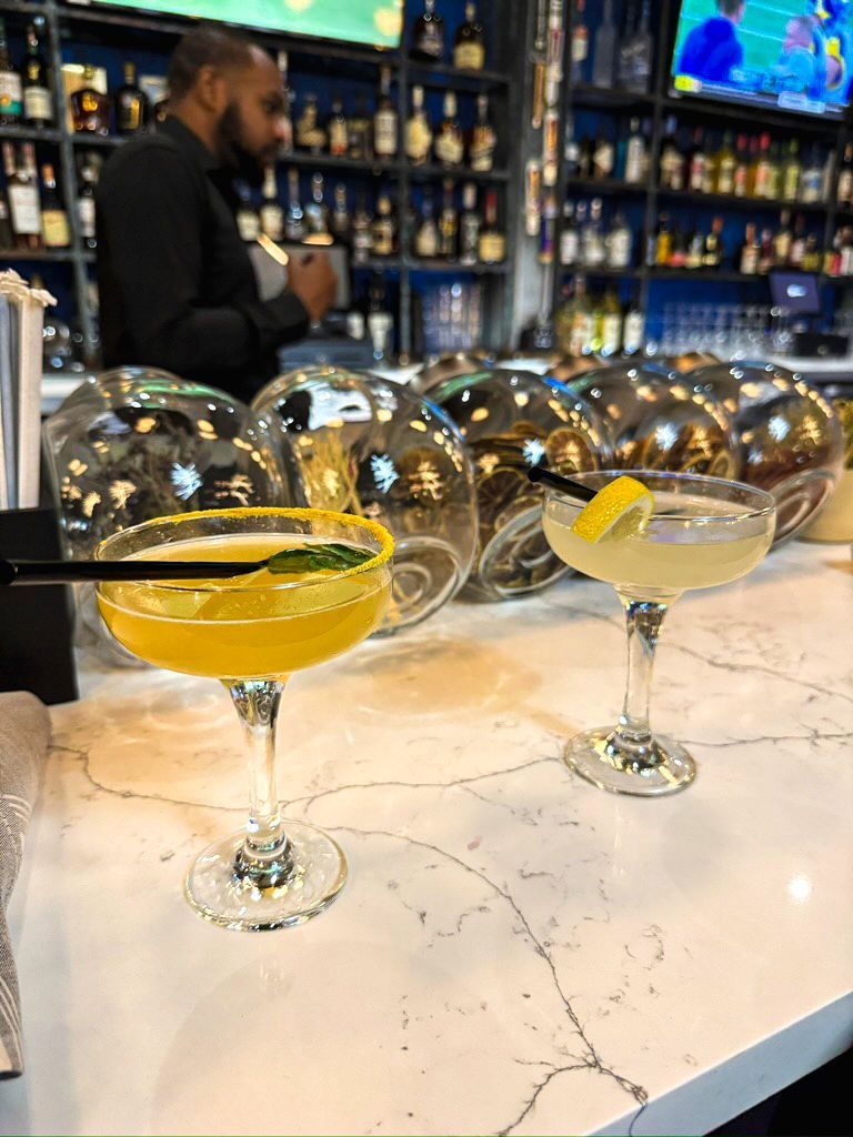 Two cocktails on a white bar, bartender in background, with glass bottles and shelves.