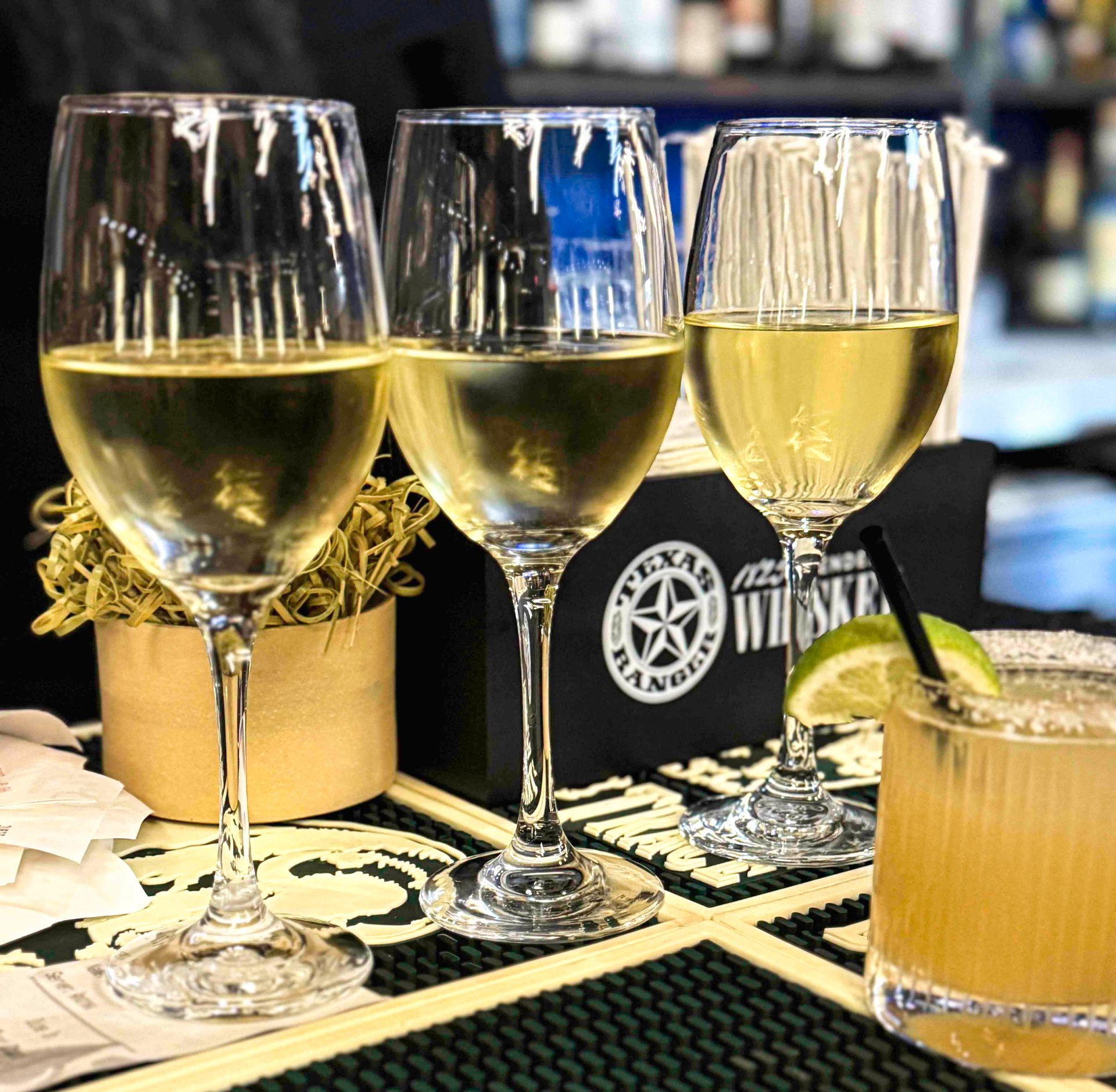 Three wine glasses and a cocktail on a bar, with a Texas Ranger whiskey box.