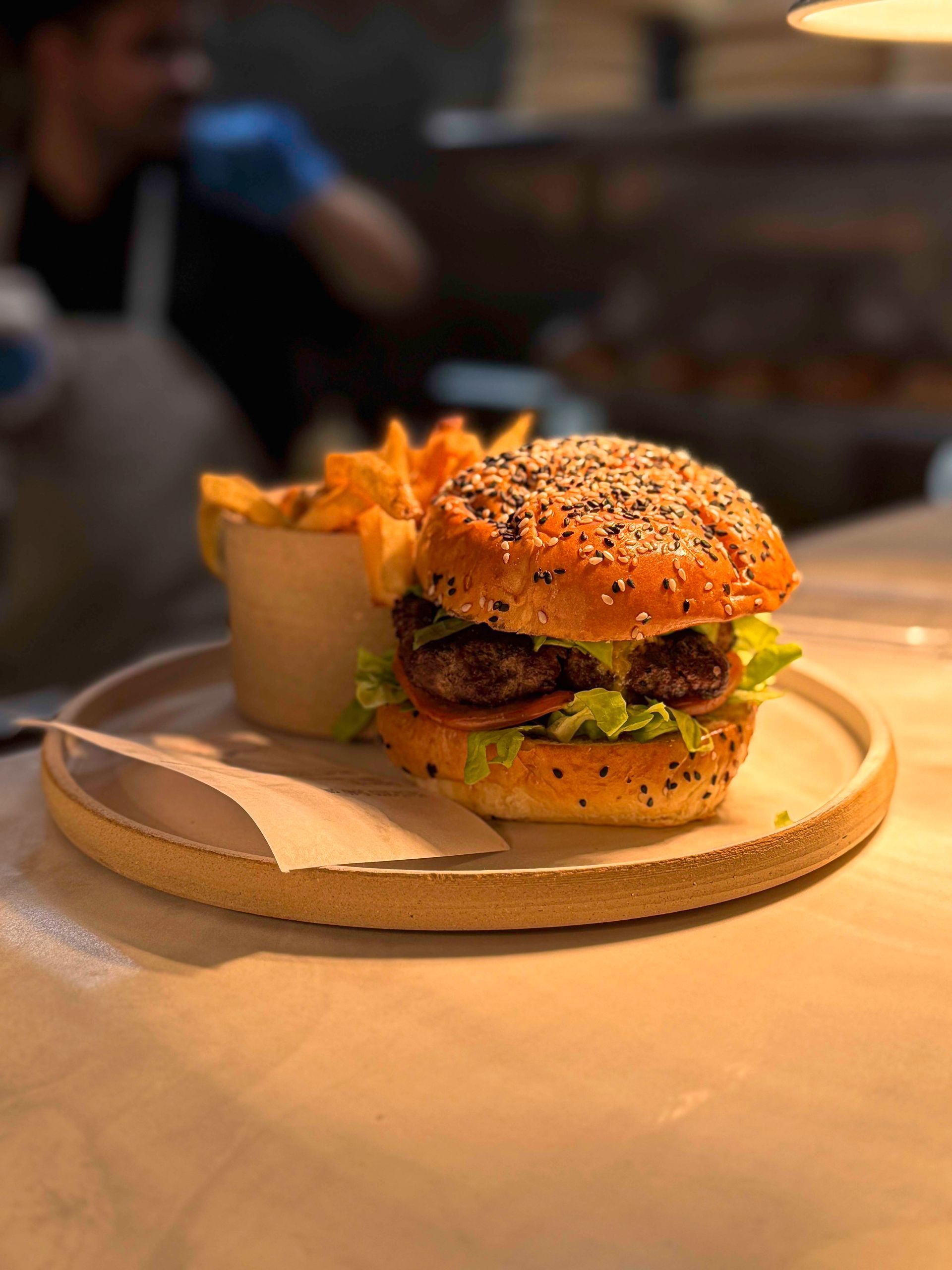 Burger with fries in a paper cup on a plate; a person in the background.