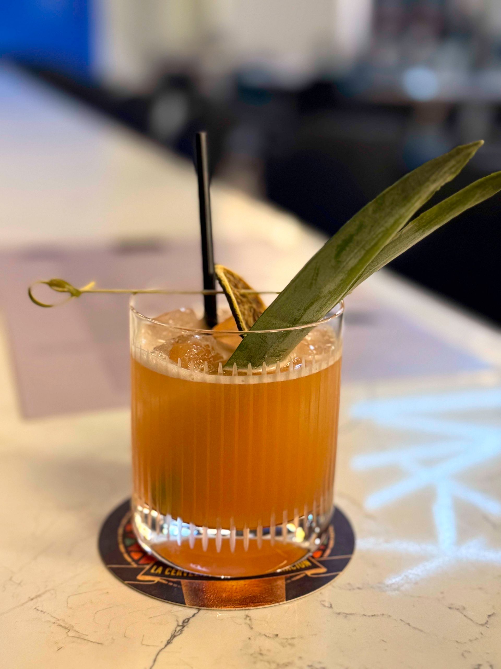 Cocktail in a ribbed glass with a pineapple leaf garnish and black straw, sitting on a coaster.