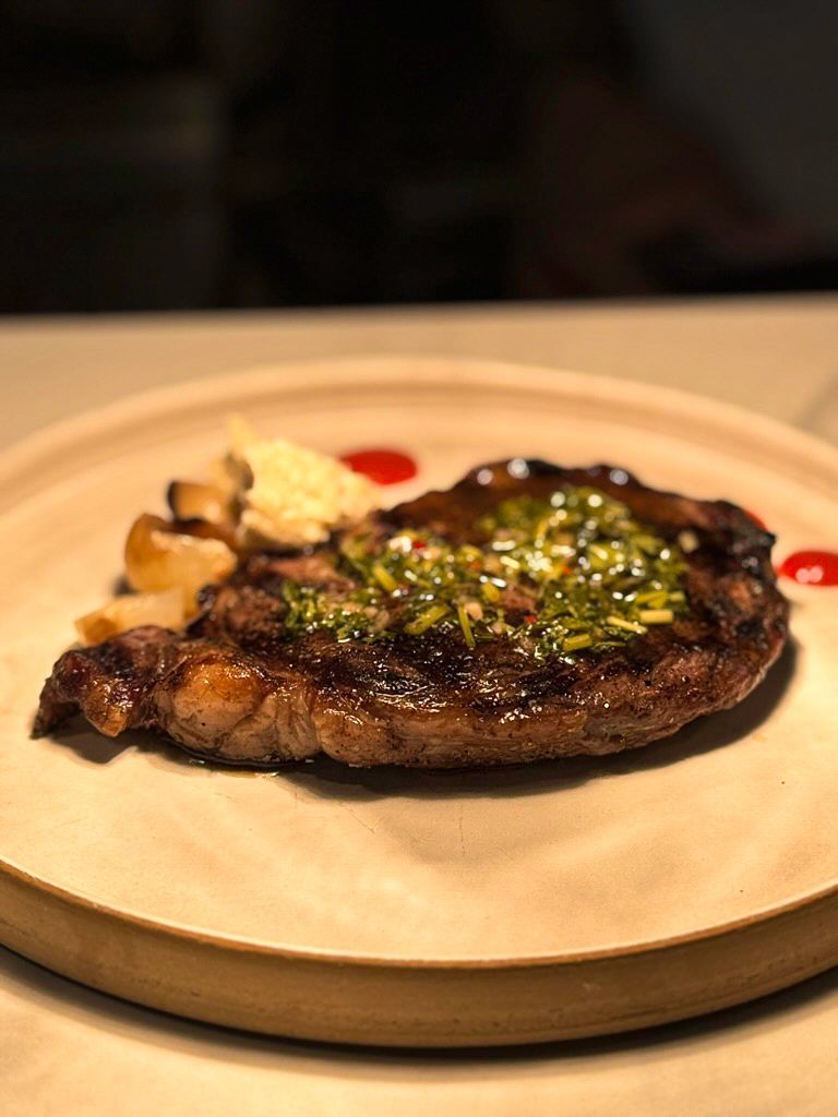 Grilled steak topped with green herbs and sauce, served with potatoes on a wooden plate.