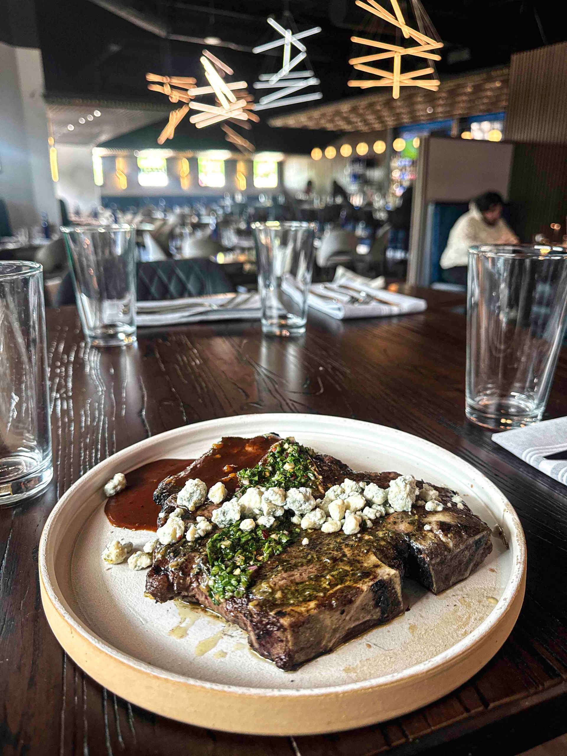 A grilled steak topped with chimichurri sauce, served with roasted garlic cloves on a minimalist ceramic plate.