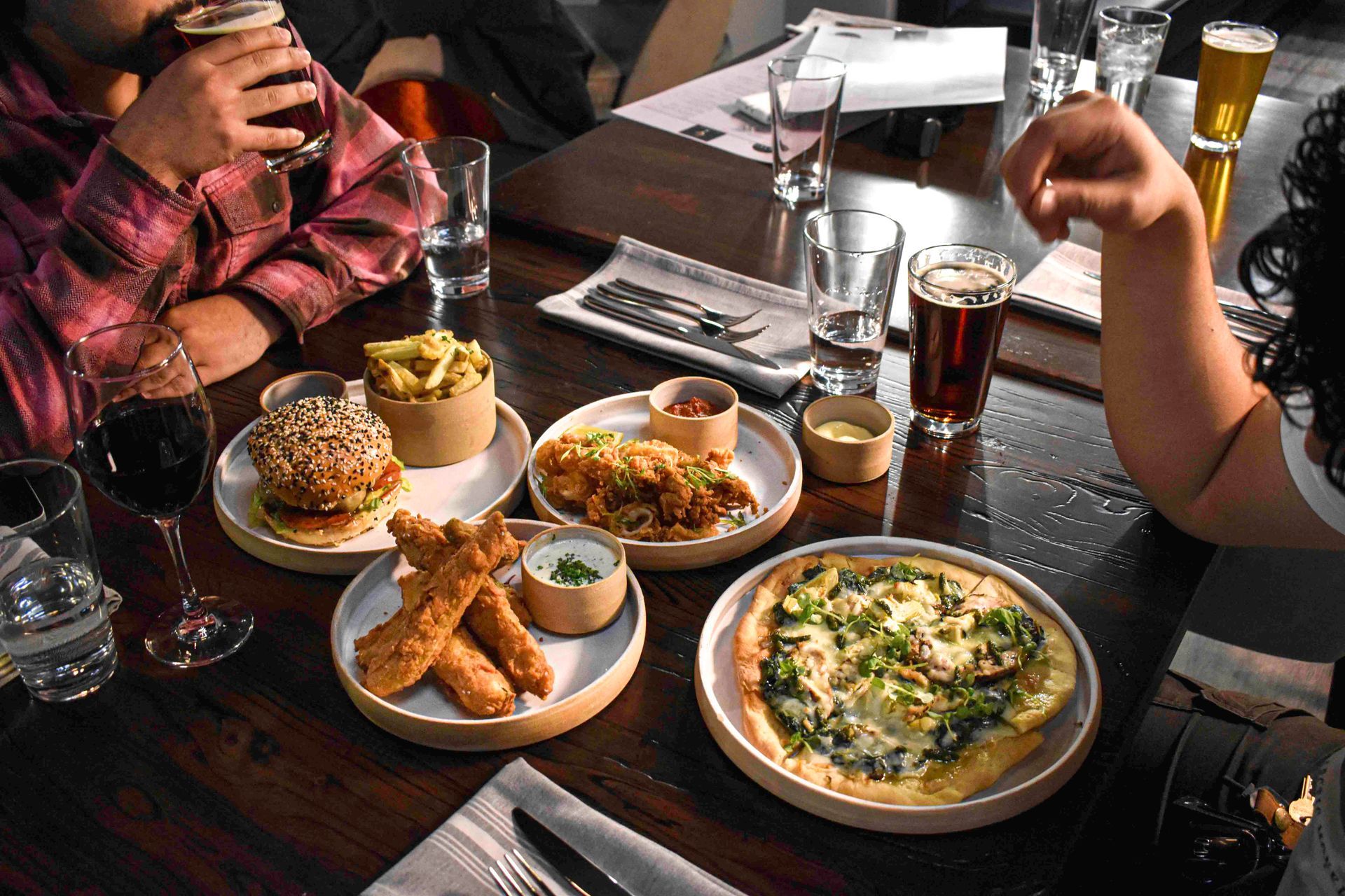 Table with food and drinks. People eating. Burger, fries, pizza, and chicken. Dark wood table.