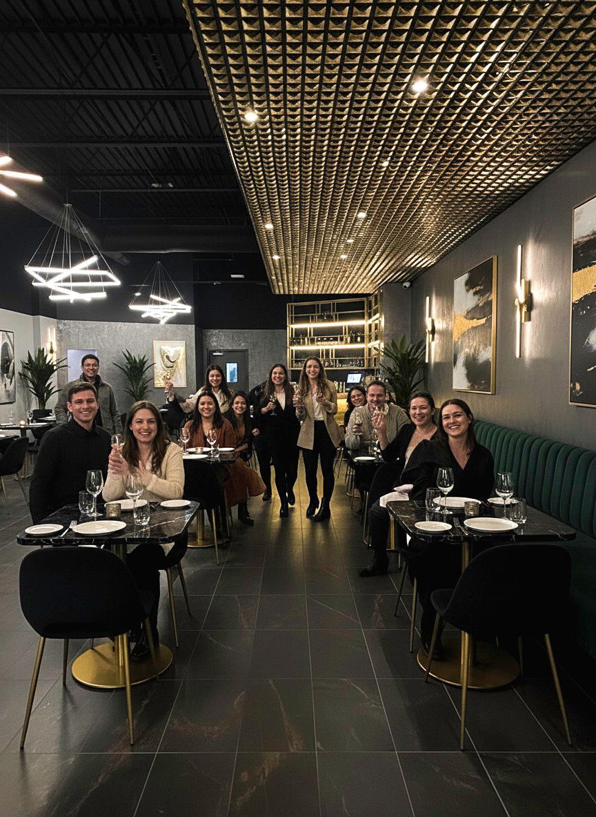 Group of people toasting at a restaurant, seated and standing. The interior has modern decor and low lighting.