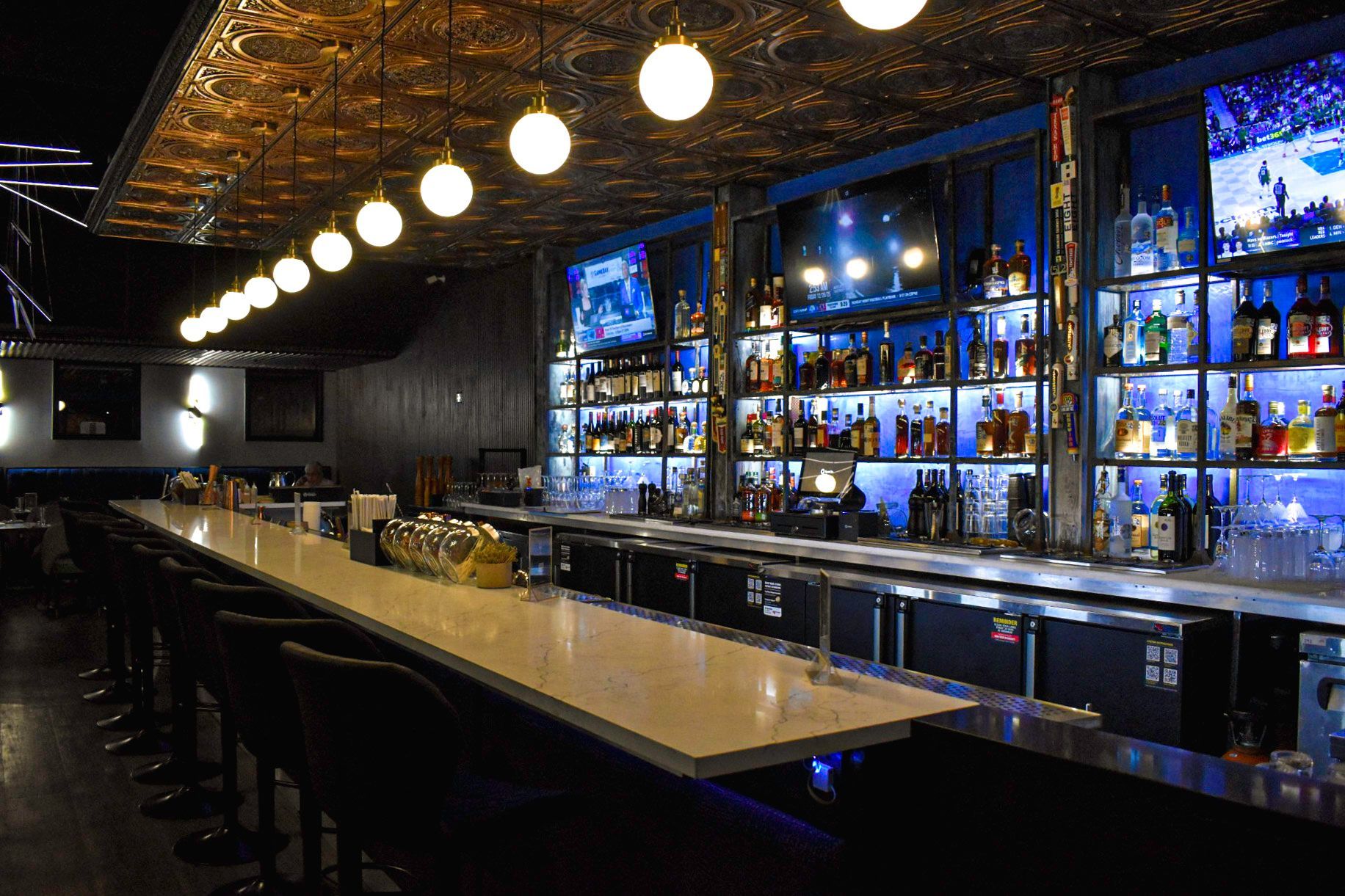Interior of a bar with a long counter, stools, shelves of liquor, and TVs. Overhead lights illuminate the space.