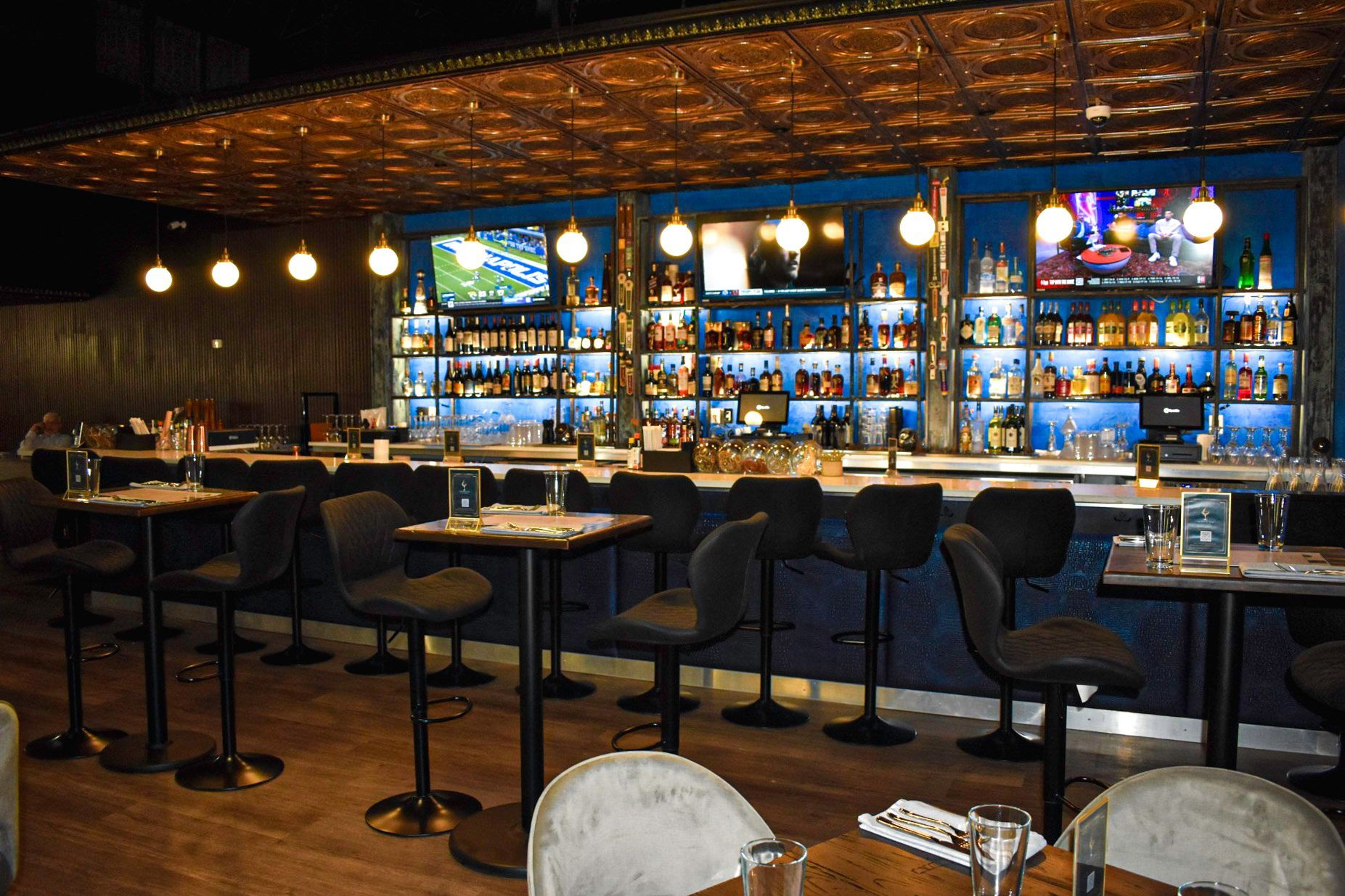 Bar with stools, tables, and a backlit liquor display. Copper-tiled ceiling, blue walls, and lit televisions.