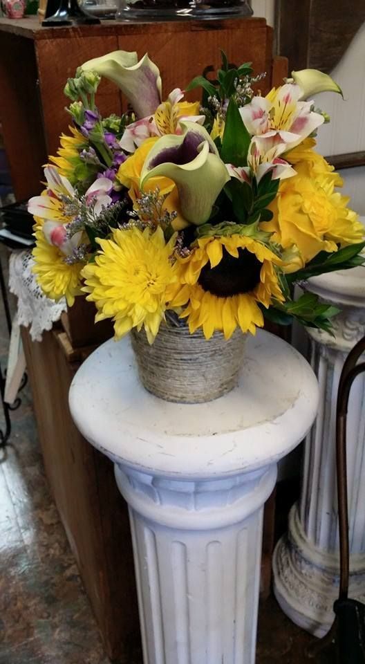 Colorful floral arrangement with yellow sunflowers and lilies in a woven vase on a white pedestal table