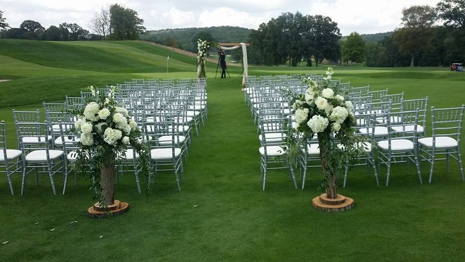 Outdoor wedding ceremony setup on a green lawn with white chairs and floral arrangements leading to an arch.