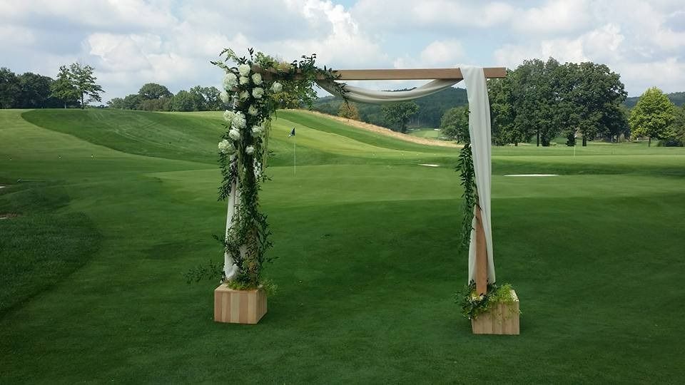Floral wedding arch with white drapes on a green lawn, framed by rolling hills and trees