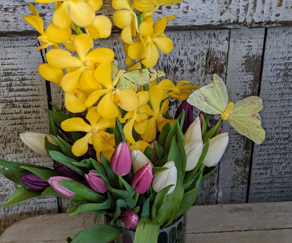 Yellow and pink flowers in a glass vase with two yellow butterflies on a rustic wooden backdrop