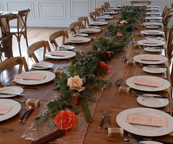 Long wooden dining table set with plates, napkins, and floral greenery centerpiece in a bright room
