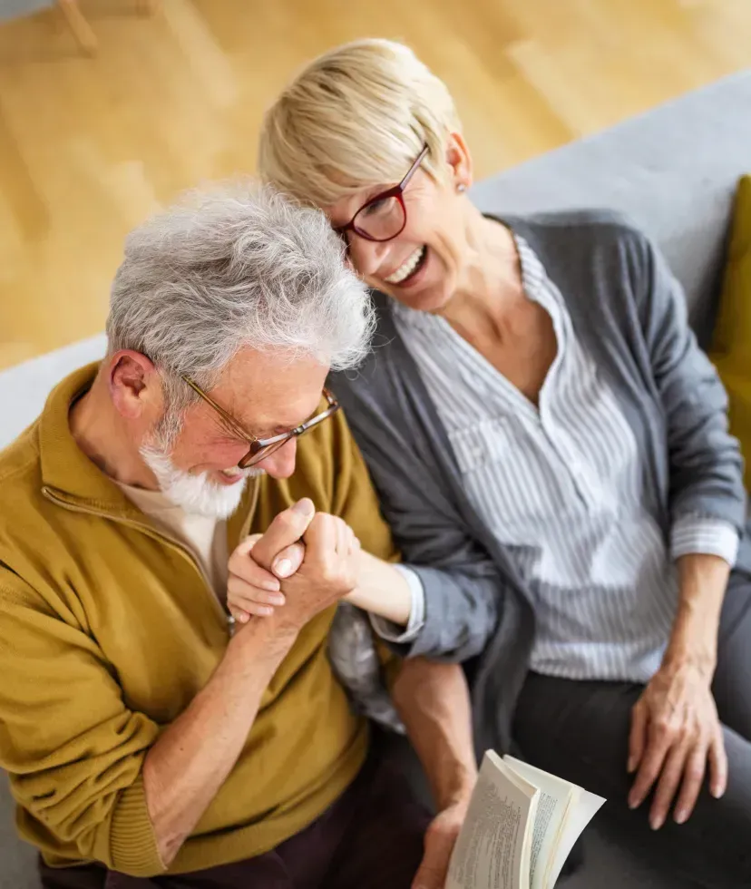 Older couple laughing, seated on couch; woman leans on man who kisses her hand, holding a book.