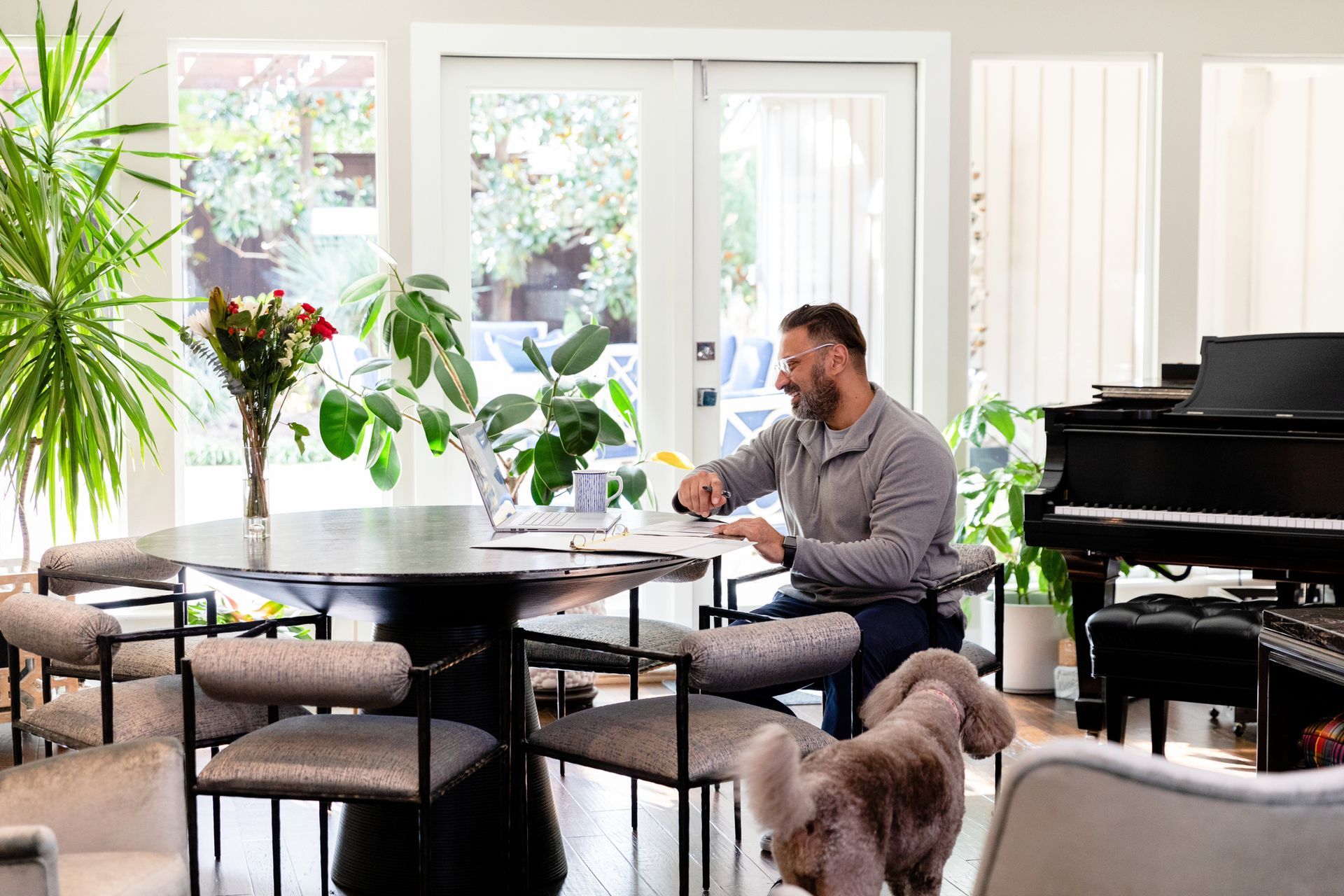 Man working at a round table with a laptop, dog nearby, and a piano in a bright room.