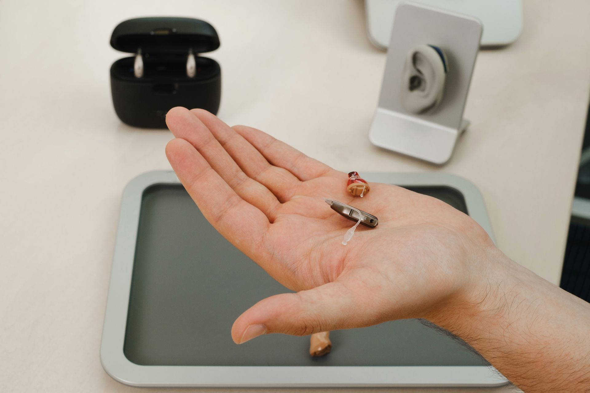 Hand holding small hearing aids, with charging case and ear mold in background.