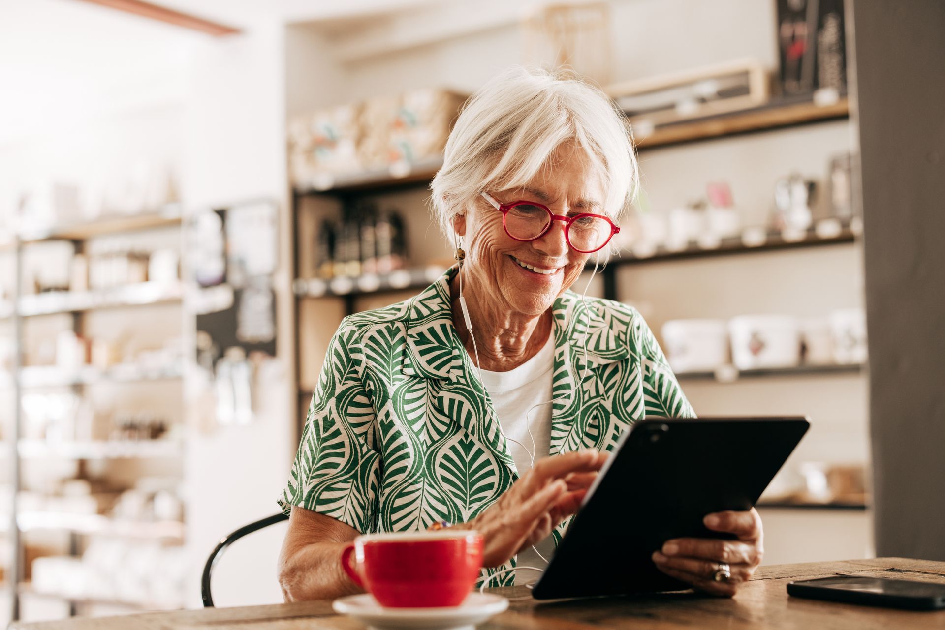 Woman with red glasses smiles, using a tablet at a cafe table with a red coffee cup.