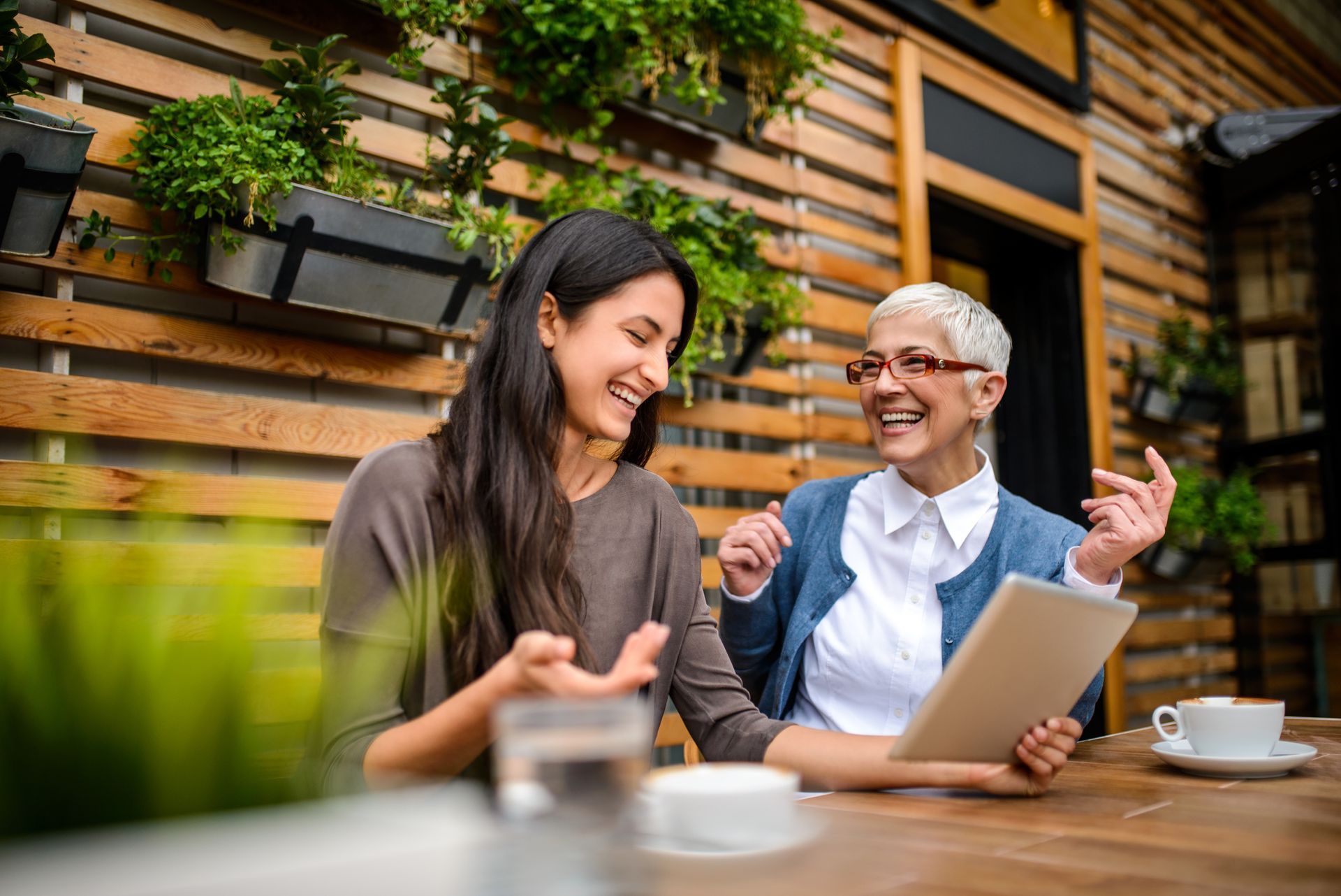 Two women laughing, looking at tablet outdoors, coffee cups, wooden wall, plants.