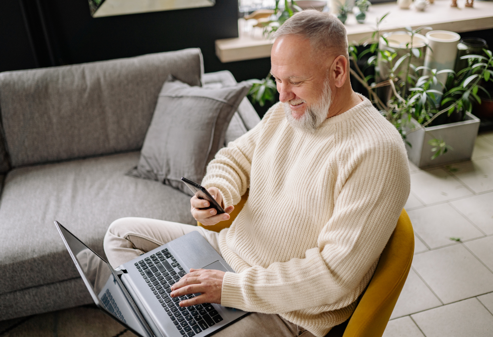 Man using laptop and phone, sitting in a chair near a couch, indoors. He is smiling.