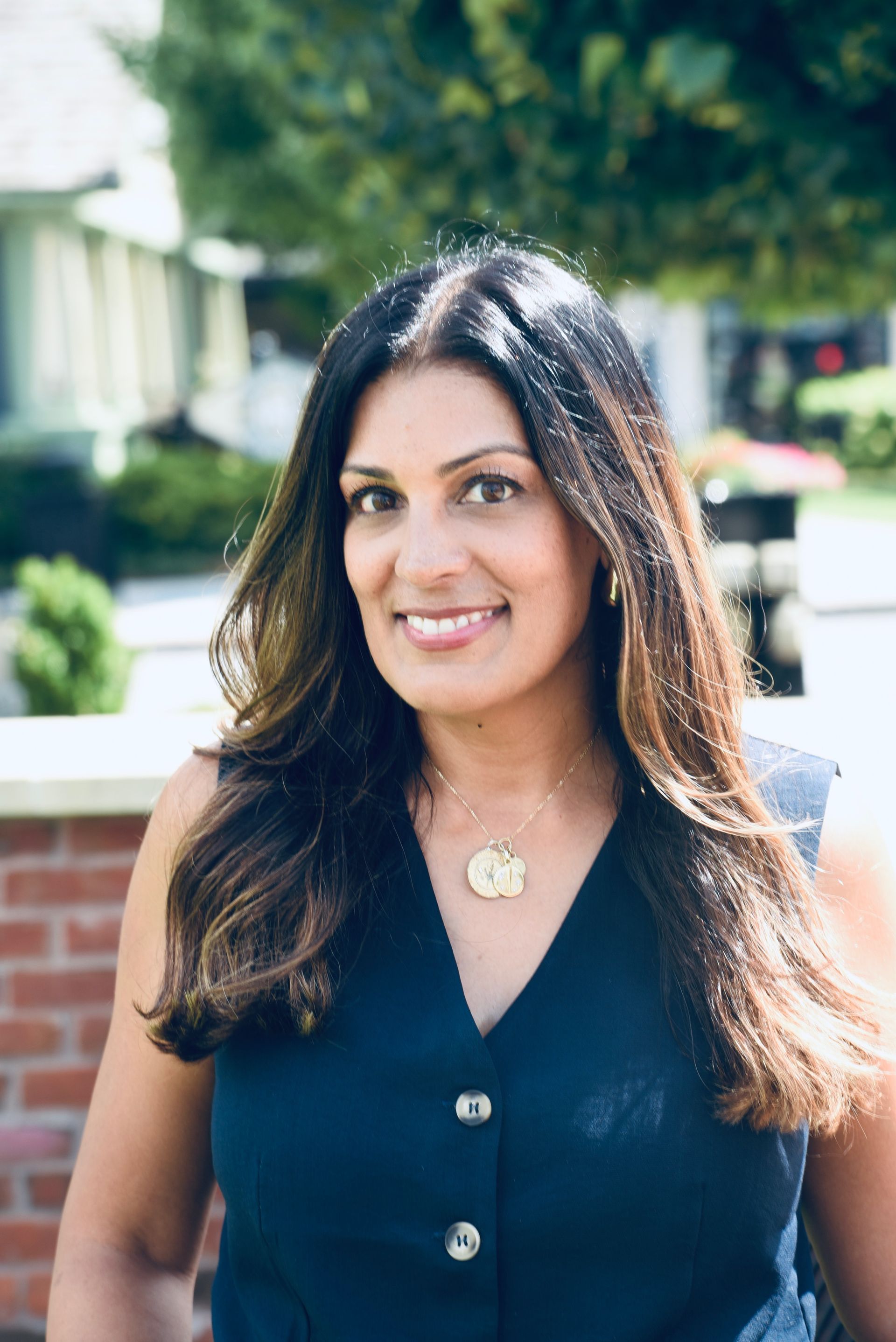 Woman with long dark hair and a navy blue top smiles outdoors in front of a brick building.