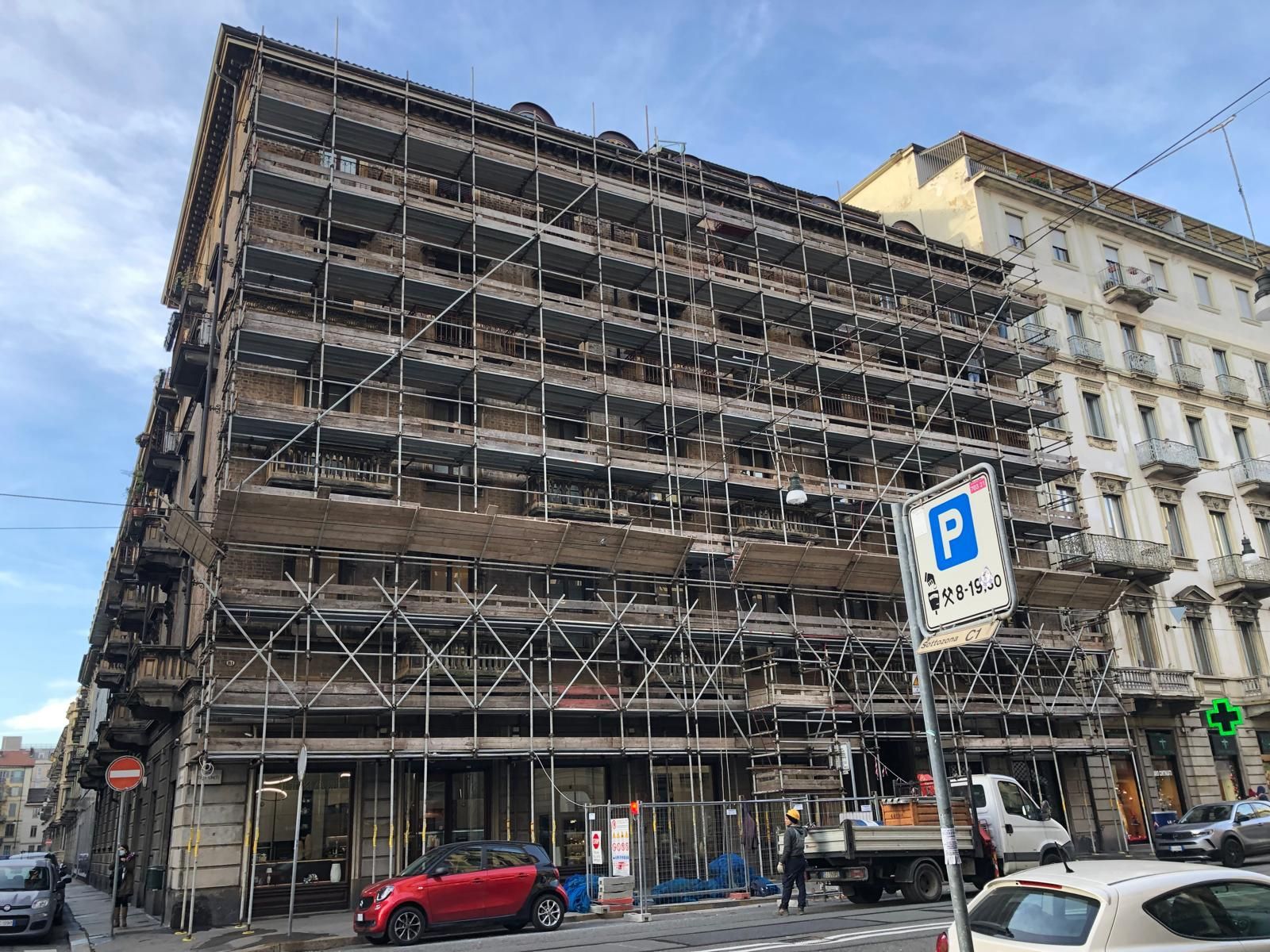 Un grande edificio è in fase di ristrutturazione con impalcature e auto parcheggiate davanti.
