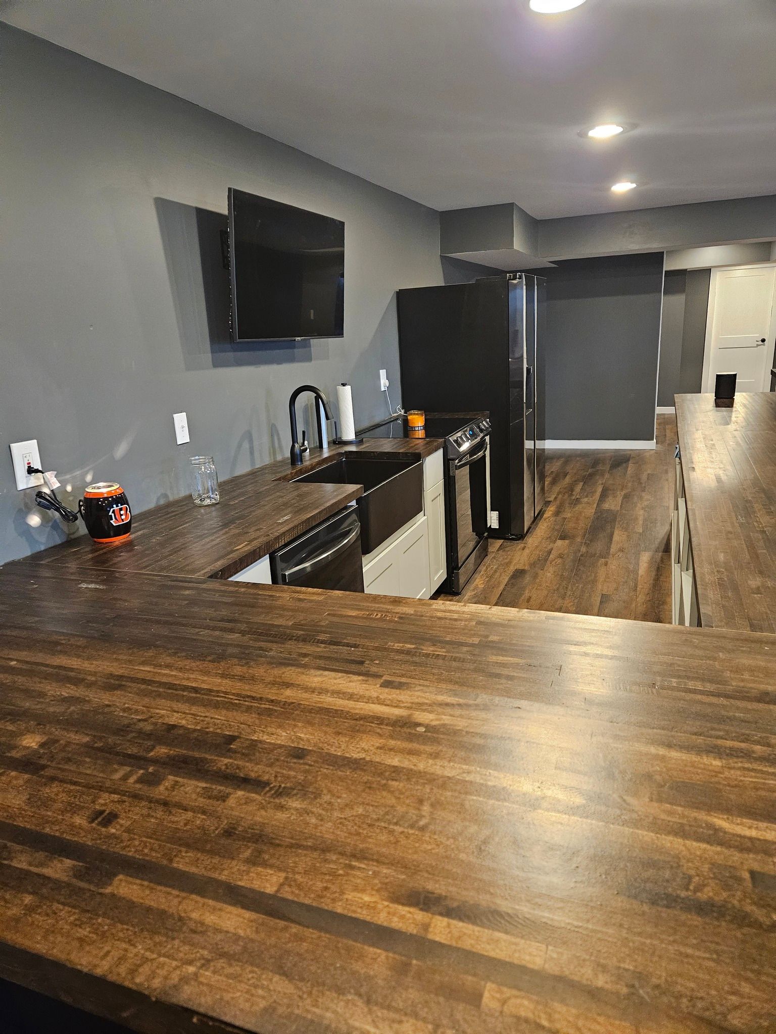 A kitchen with dark brown countertops, a black fridge, and a TV mounted on a gray wall.