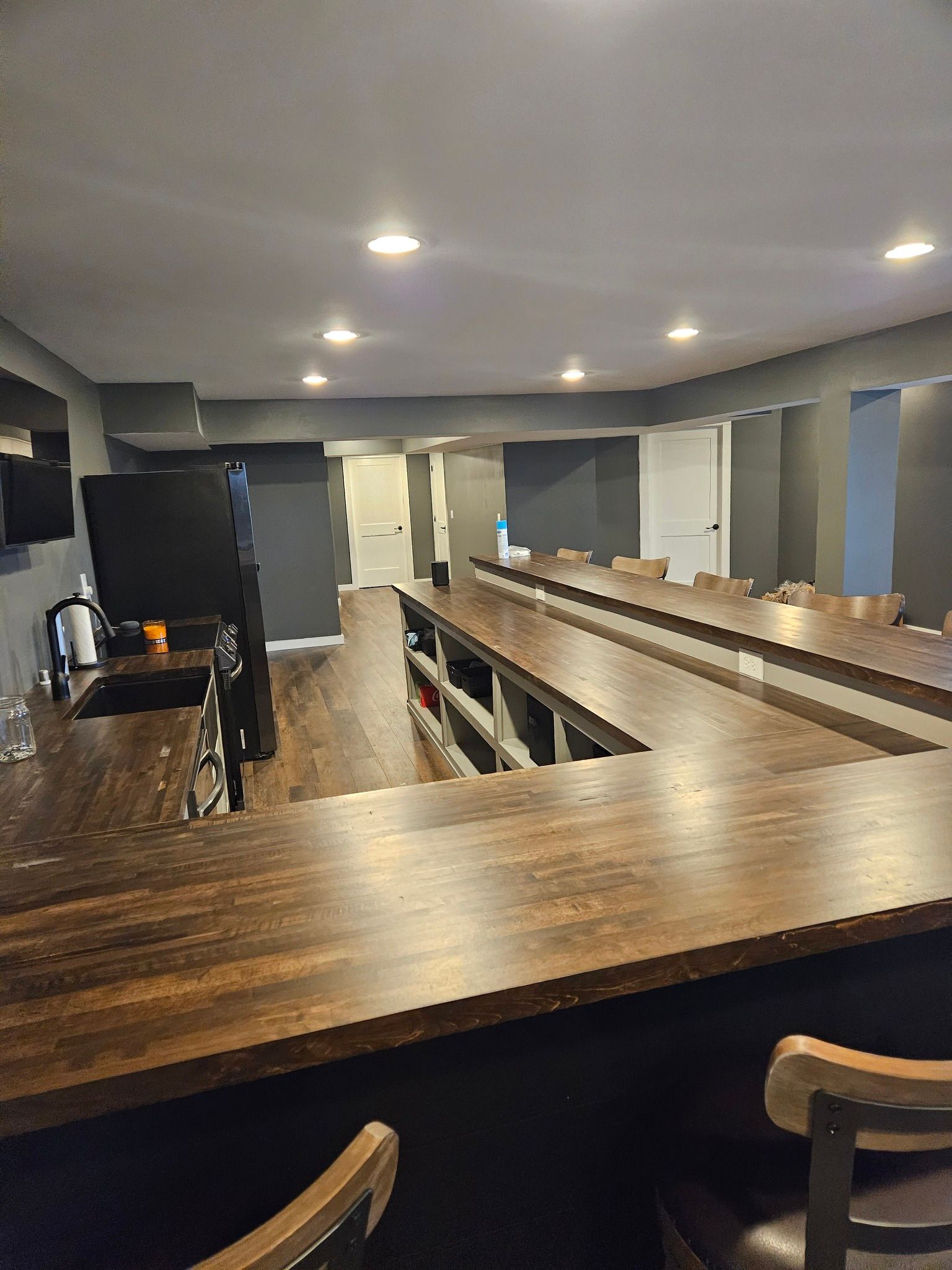 U-shaped wooden bar in a dimly lit room with gray walls and recessed lighting. Bar stools in the foreground.