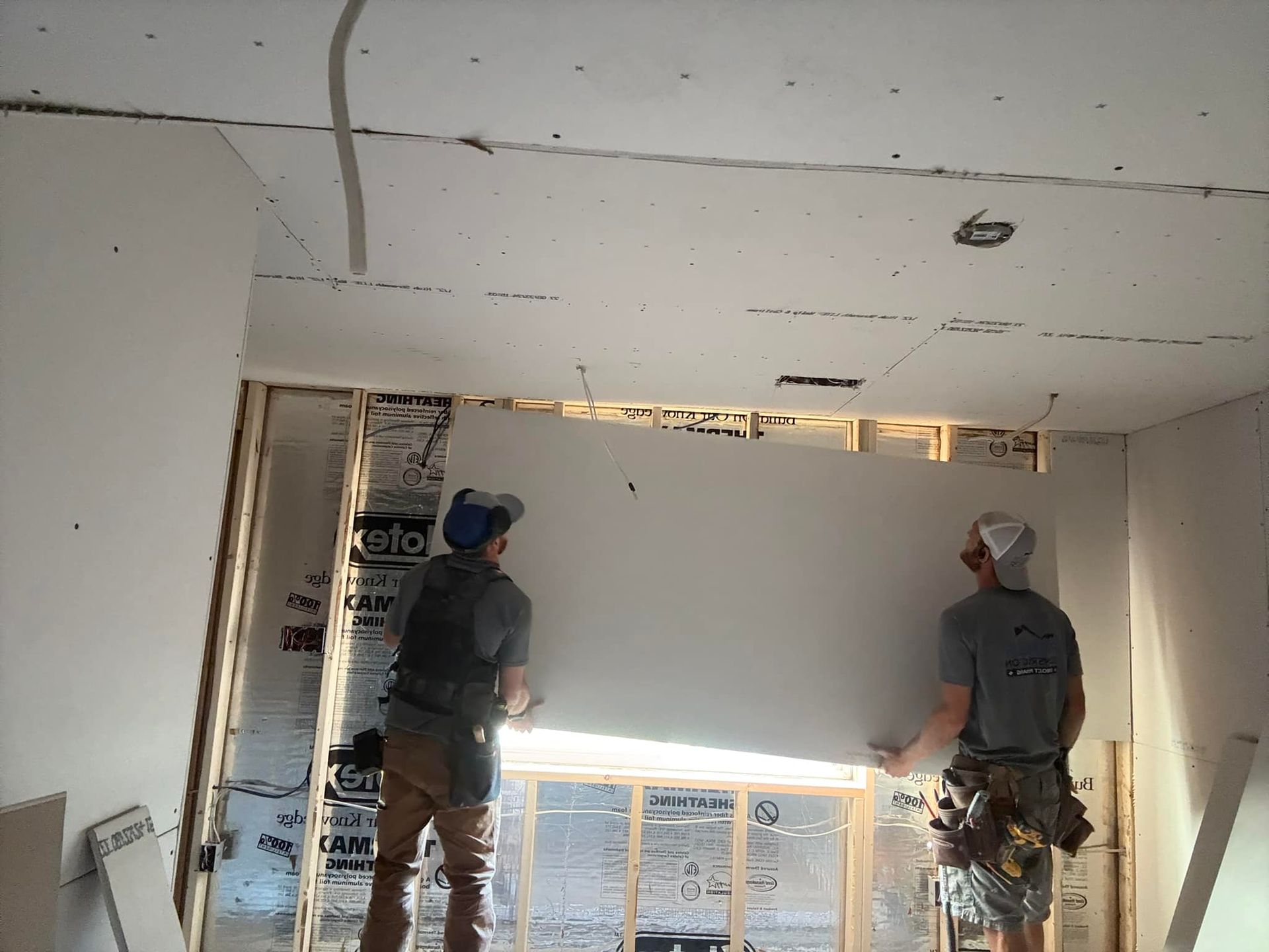 Two construction workers installing drywall on a room's ceiling and wall during a home renovation.