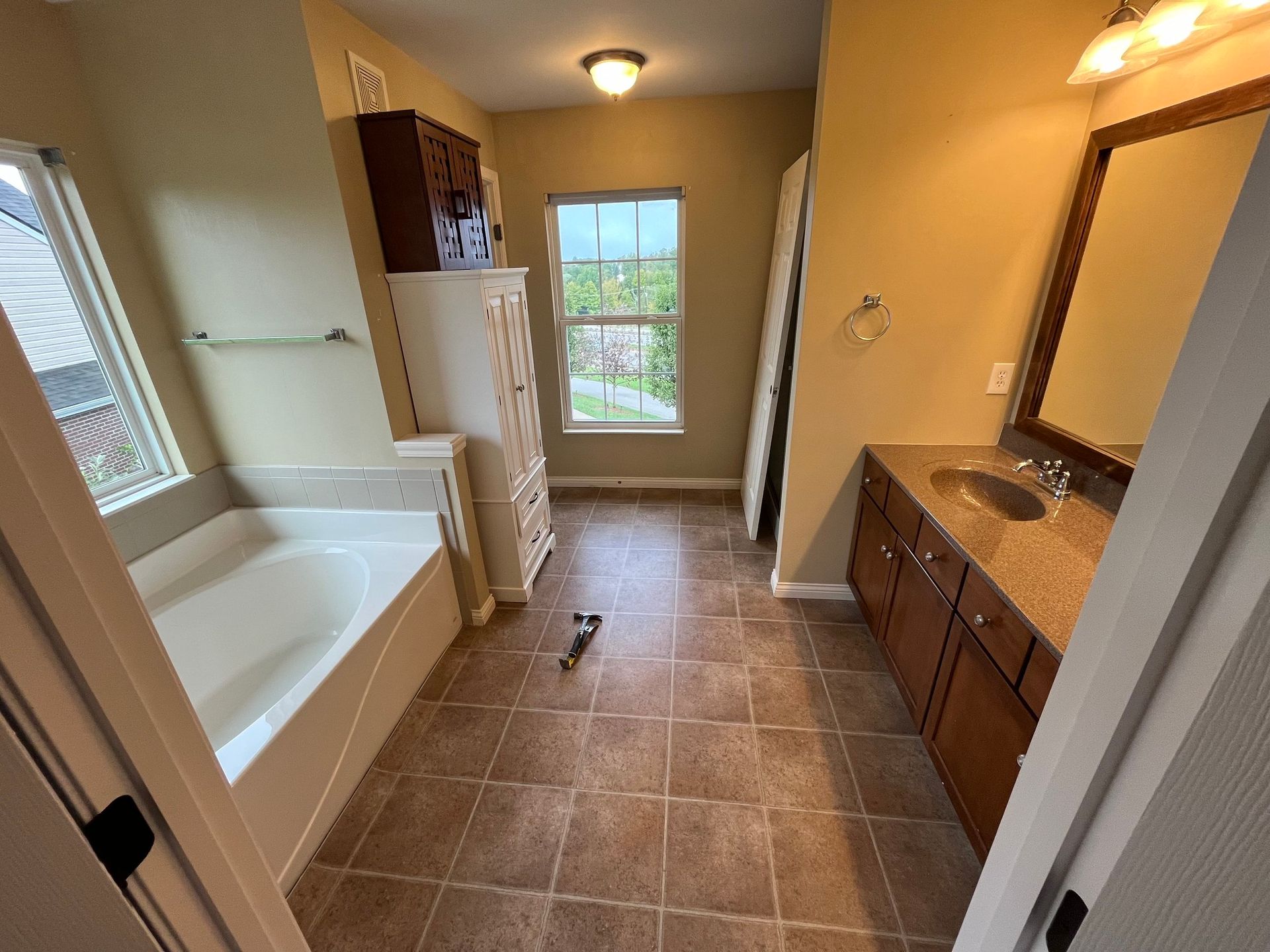 Bathroom with tub, vanity, and window. Brown floor, beige walls.  Brown cabinets, white tub and cabinet.