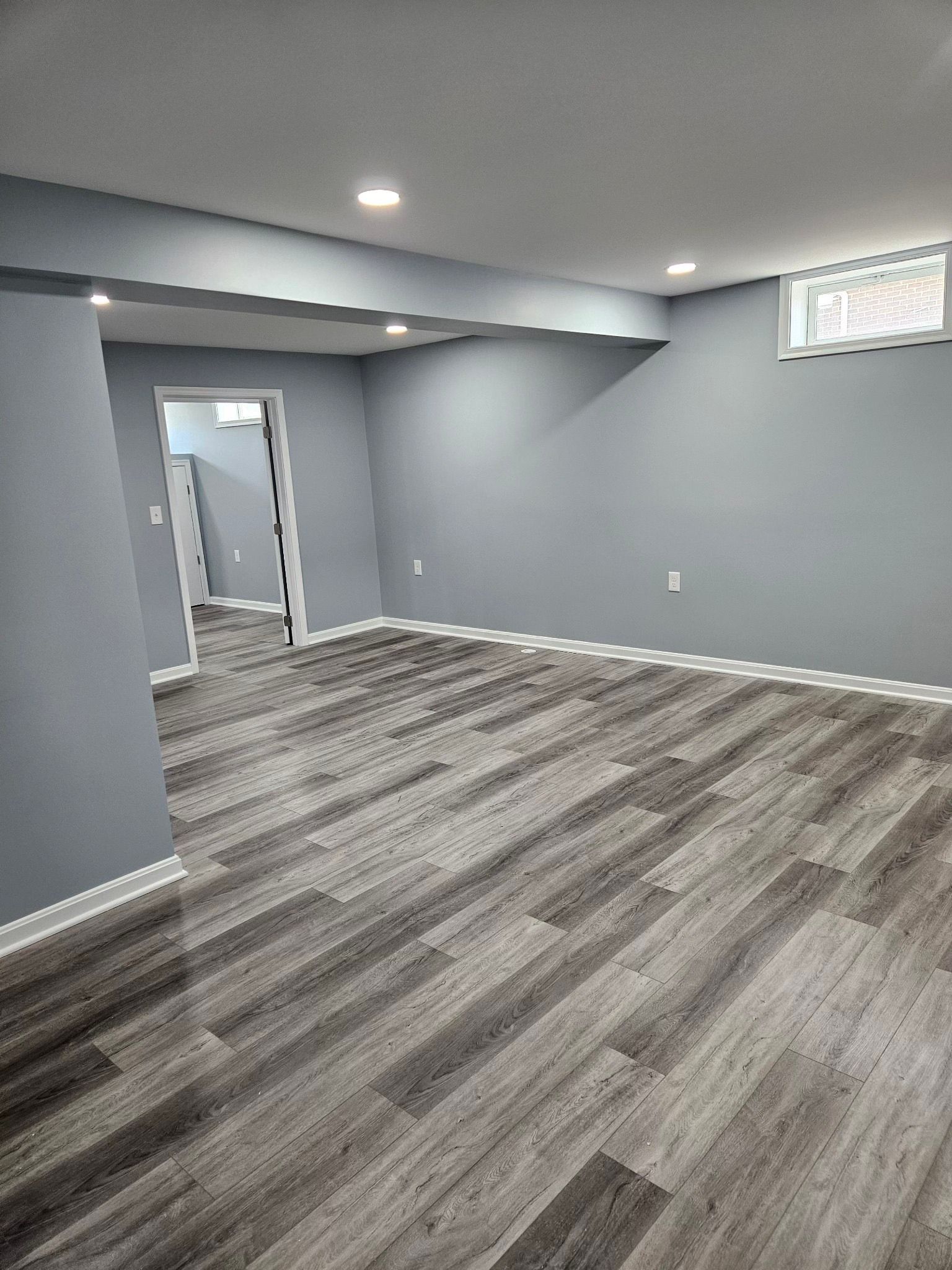 Empty basement room with grey walls, wood-look flooring, white trim, and recessed lighting.
