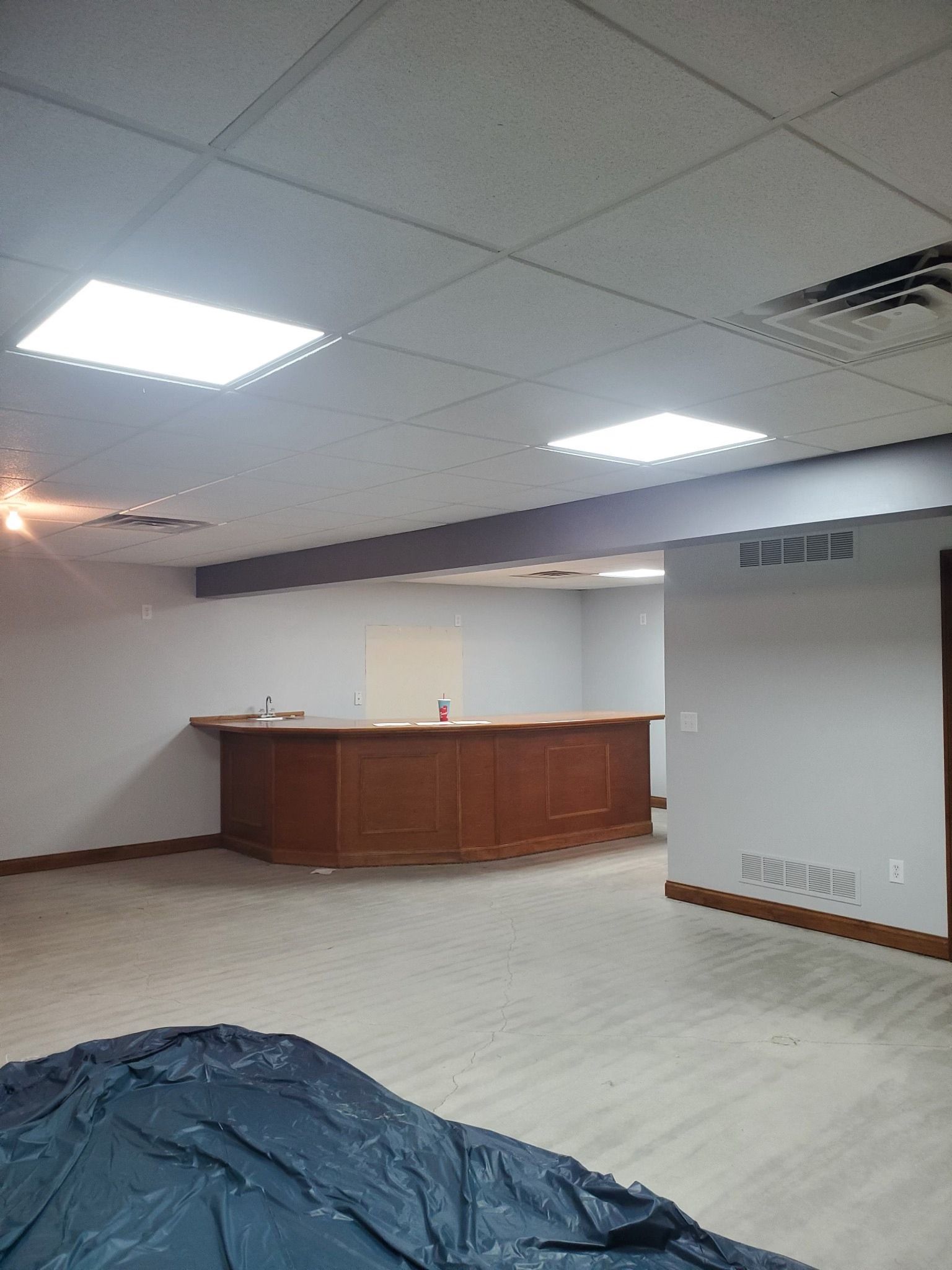 A basement bar with a wooden counter and light-colored carpet. Two ceiling lights are on.