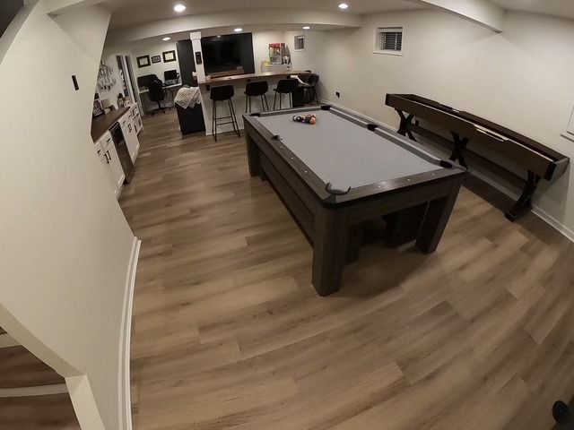 Basement recreation room with pool table, shuffleboard, and bar seating. Light wood floors and white walls.