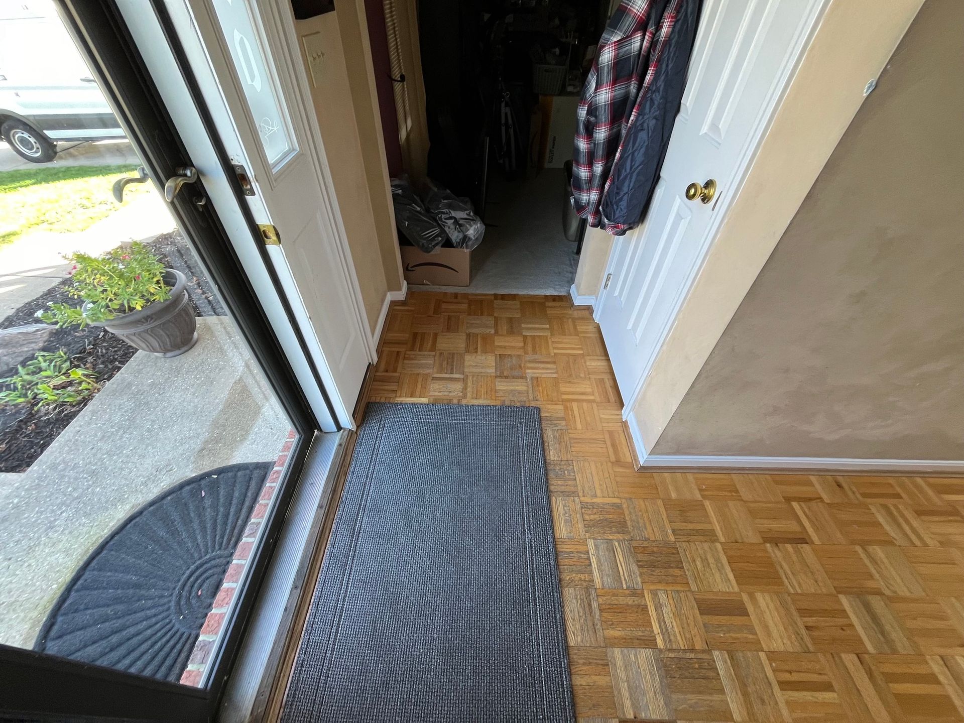 Entryway: open door, gray doormats on wood floor, coat hanging on door, plants outside.