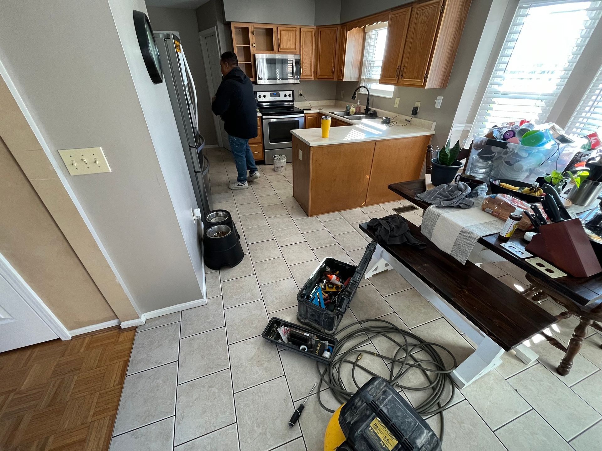 Man in kitchen with tools, cabinets, table, debris. Beige tile floor.