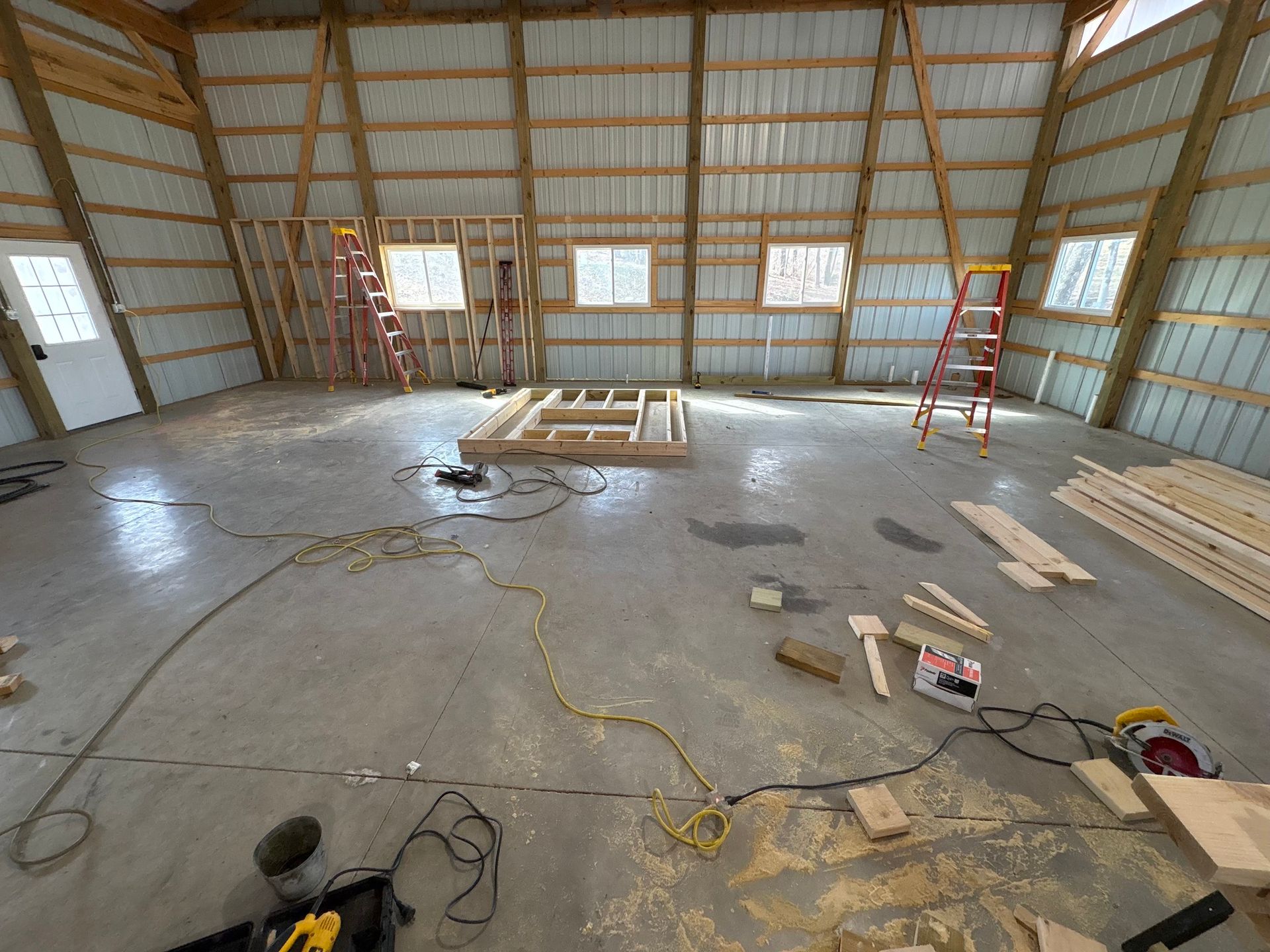 Interior of a large pole barn under construction. Concrete floor, wooden framing, windows, and ladders are visible.