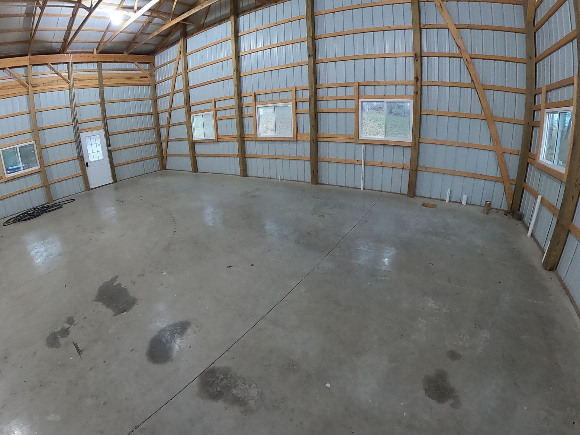 Inside of a spacious, empty gray concrete floored barn with windows, metal walls, and wooden beams.
