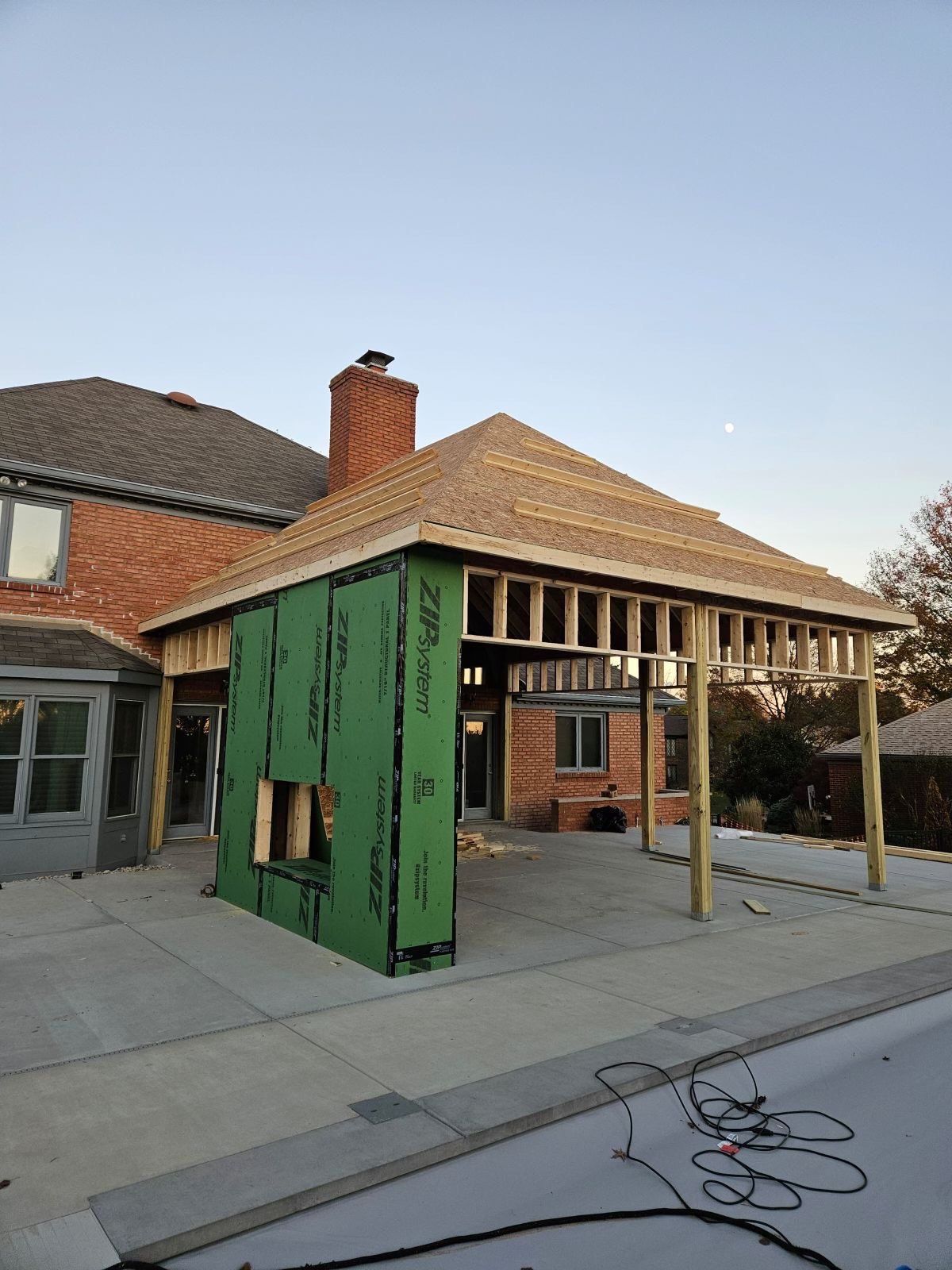 Construction of a patio cover attached to a brick house with exposed framing, green sheathing, and a pool in the foreground.